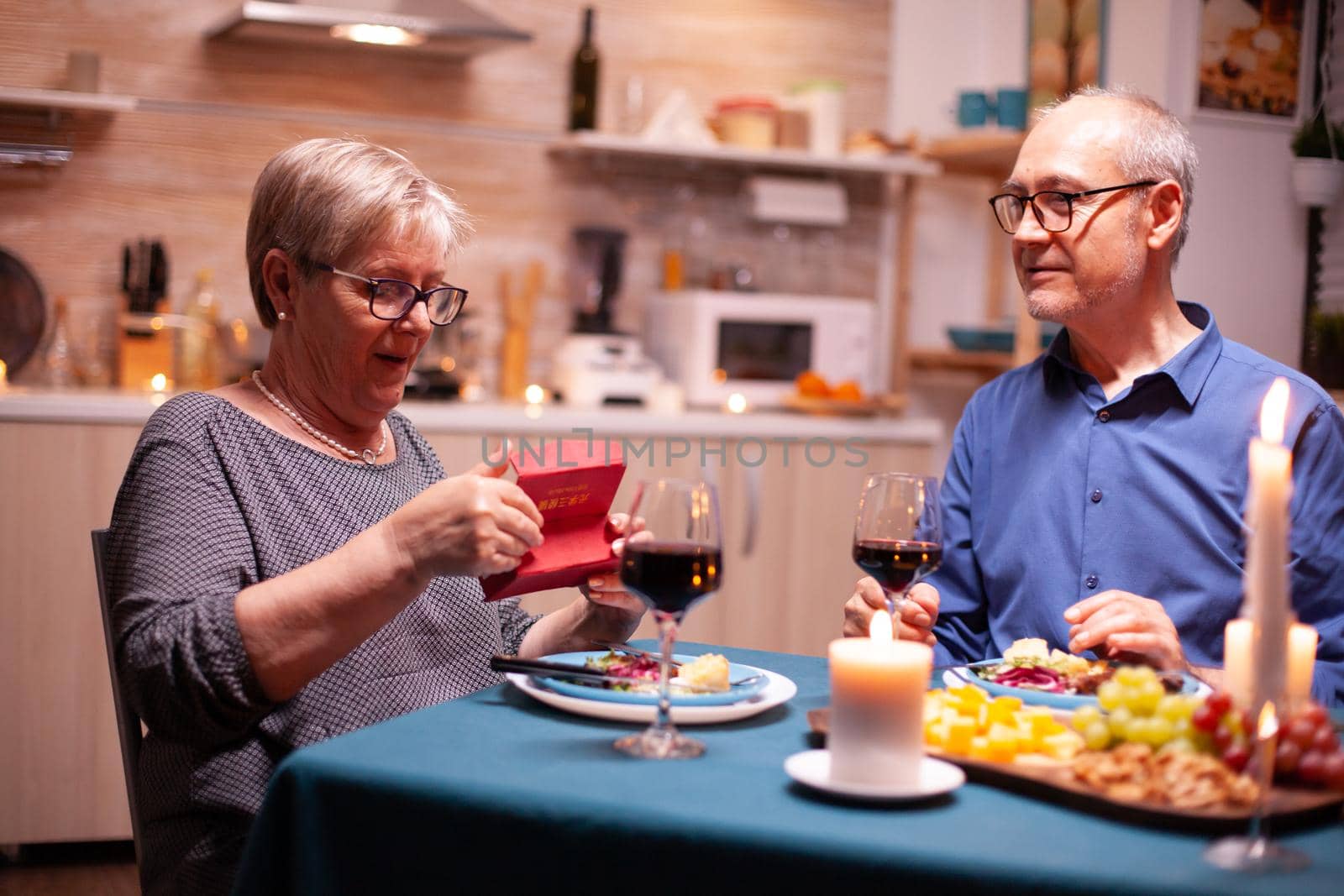 Senior woman looking surprised at gift box from husband during dinner in kitchen. Happy cheerful elderly couple dining together at home, enjoying the meal, celebrating their marriage , surprise holiday.