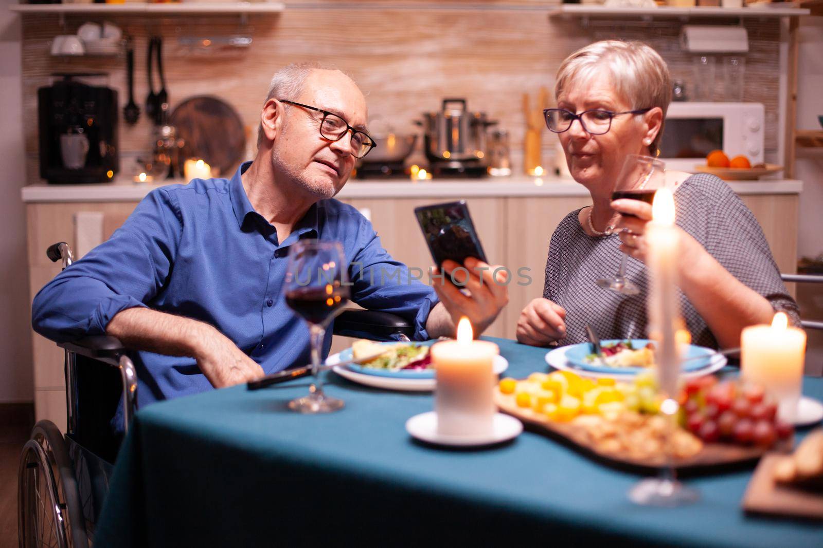 Man in wheelchair holding phone while having dinner with wife in kitchen. Scrolling and showing photos. Imobilized handicapped senior husband scolling on phone enjoying the festive meal.