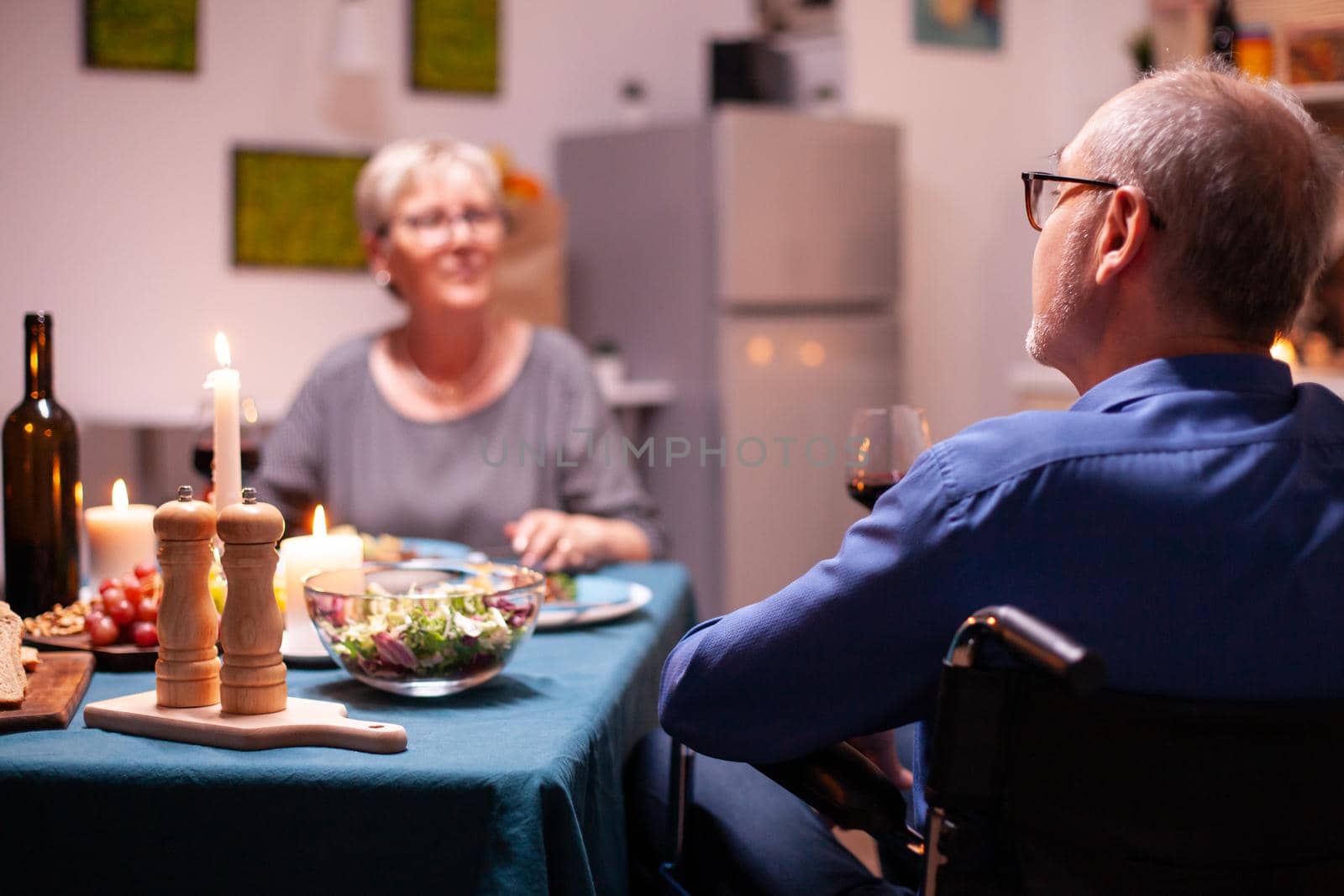 Handicapped man holding whine glass sitting in wheelchair during festive dinner. Happy cheerful senior elderly couple dining together in the cozy kitchen, enjoying the meal, celebrating their anniversary.