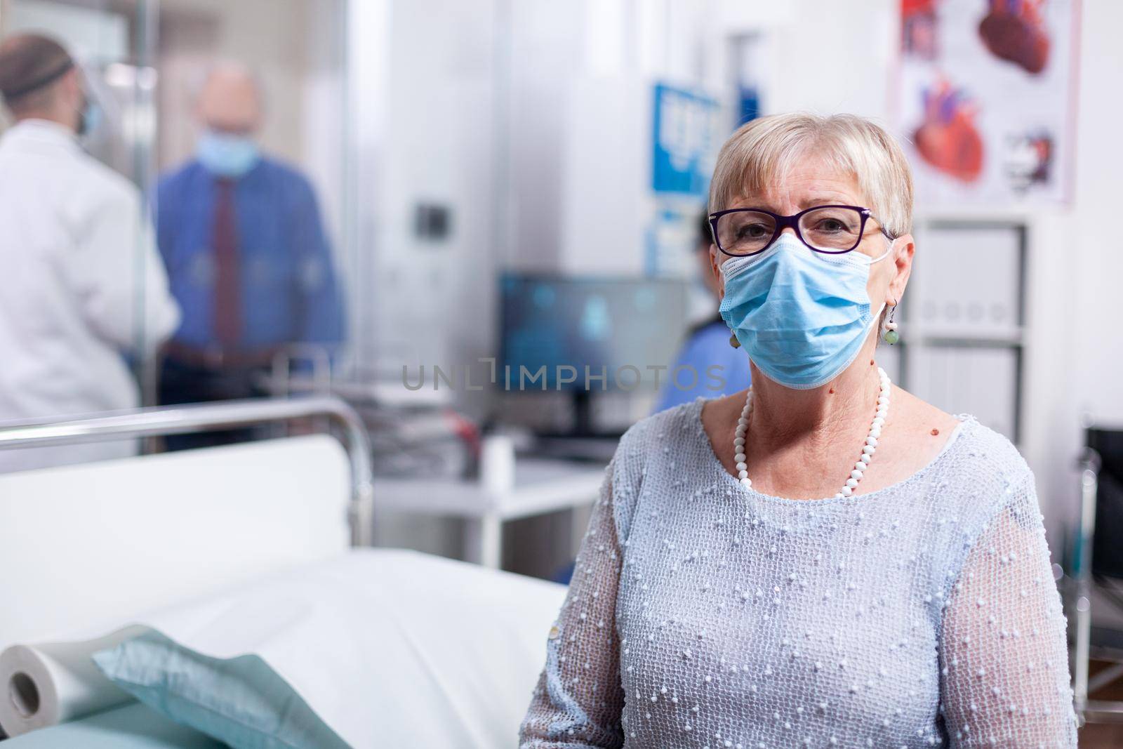 Elderly sick patient with face mask in hospital cabinet waiting for doctor. Global health crisis, medical system during pandemic, sick elderly patient in private clinic.