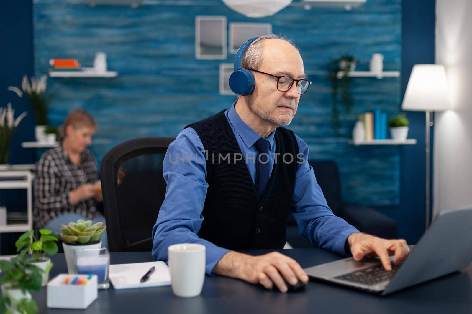 Senior businessman listening music wearing headphones working on laptop. Elderly man entrepreneur in home workplace using portable computer sitting at desk while wife is reading a book sitting on sofa.