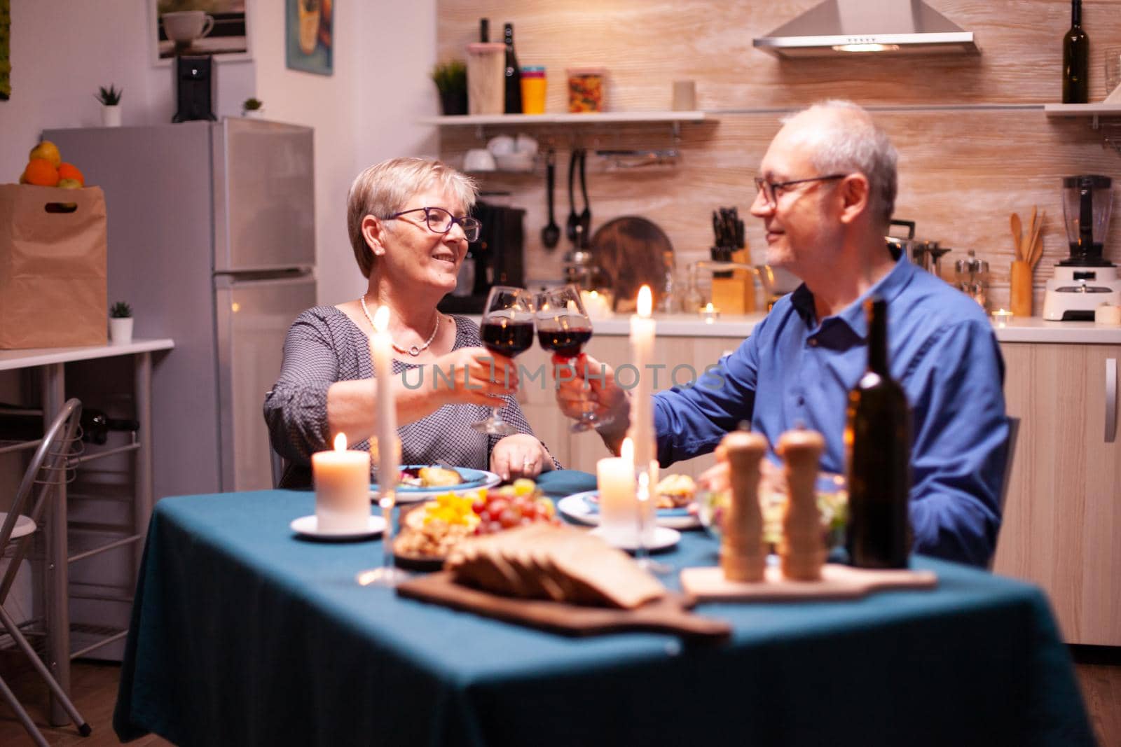 Elderly couple toasting wine glasses for festive dinner. Happy cheerful senior elderly couple dining together in the cozy kitchen, enjoying the meal, celebrating their anniversary.