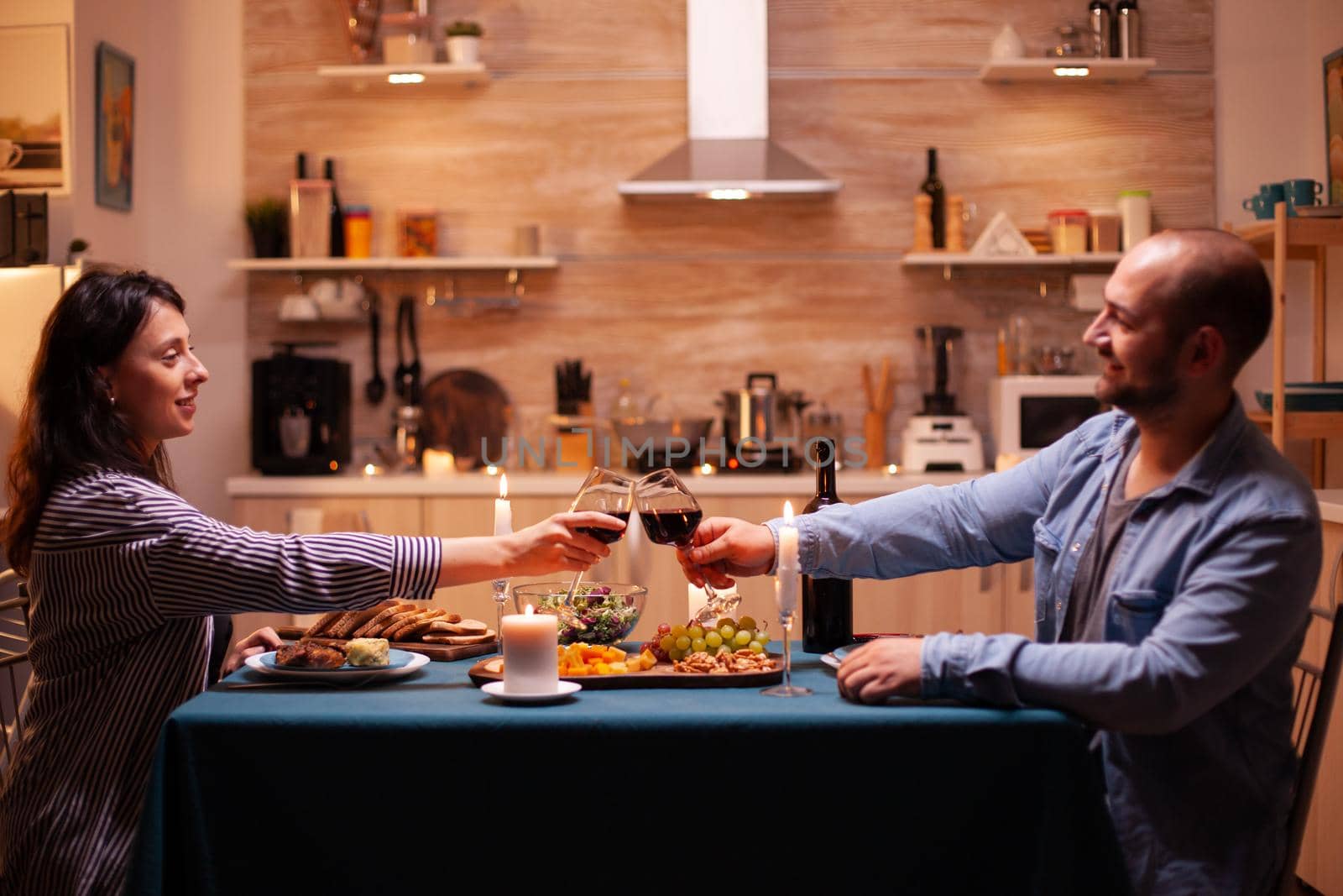 Positive husband and wife holding wine glasses in kitchen in the evening. Relax happy people clinking, sitting at table in kitchen, enjoying the meal, celebrating anniversary in the dining room.