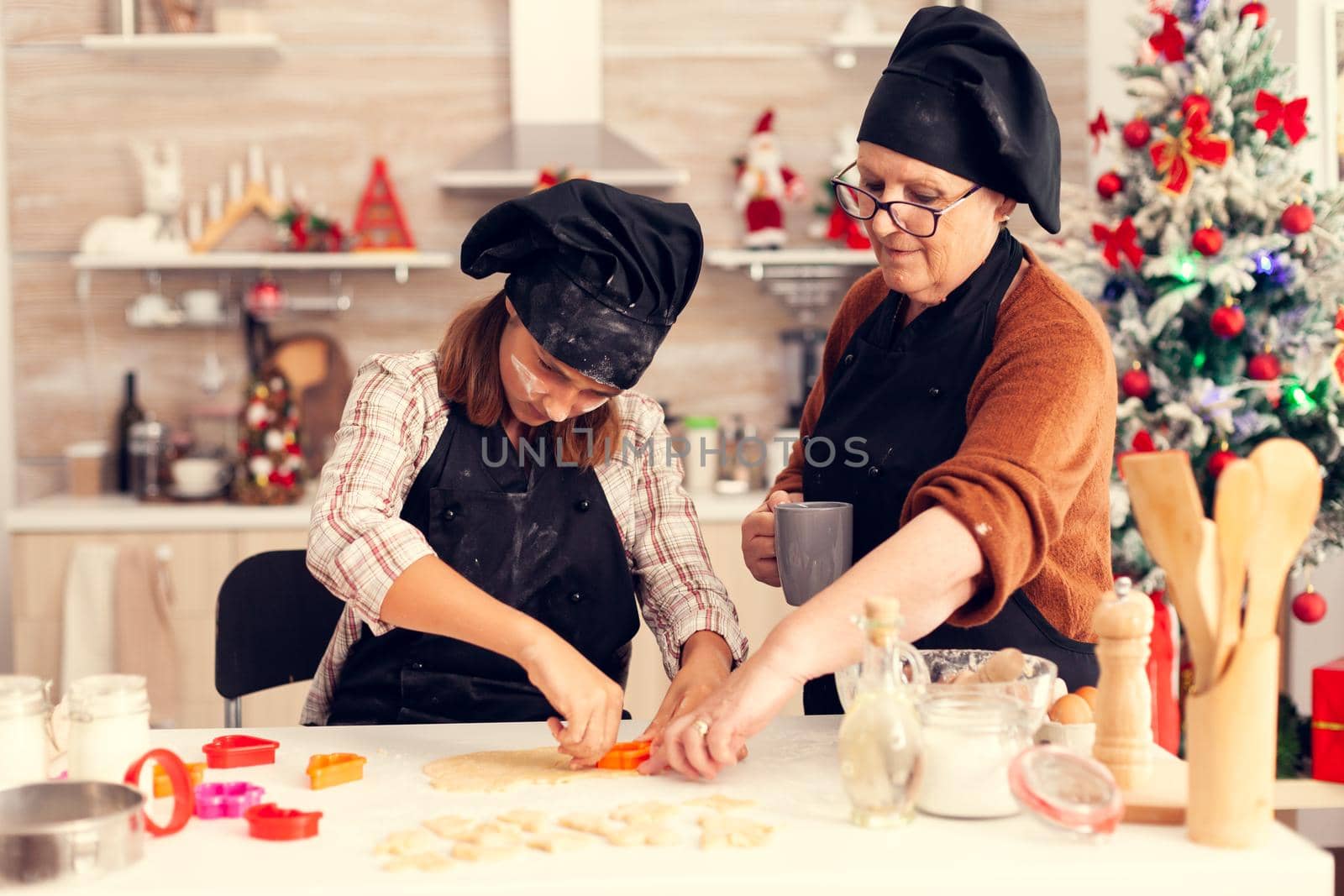 Grandchild using dough shape cutter on christmas day with help from grandmother. Happy cheerful joyfull teenage girl helping senior woman preparing sweet cookies to celebrate winter holidays.