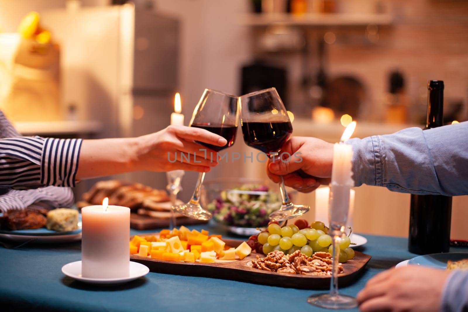 Close up of clinking glasses with wine during romantic dinner dinner celebrating relationship. Happy cheerful young couple dining together in the cozy kitchen, enjoying the meal, celebrating anniversary romantic toast