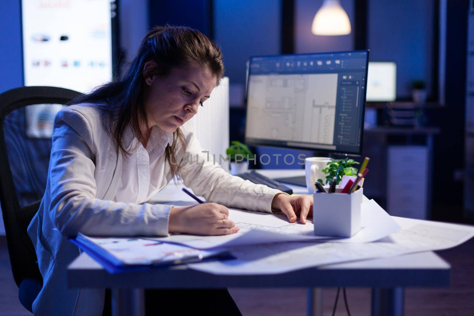 Overworked woman architect checking and matching blueprints sitting at office desk by DCStudio