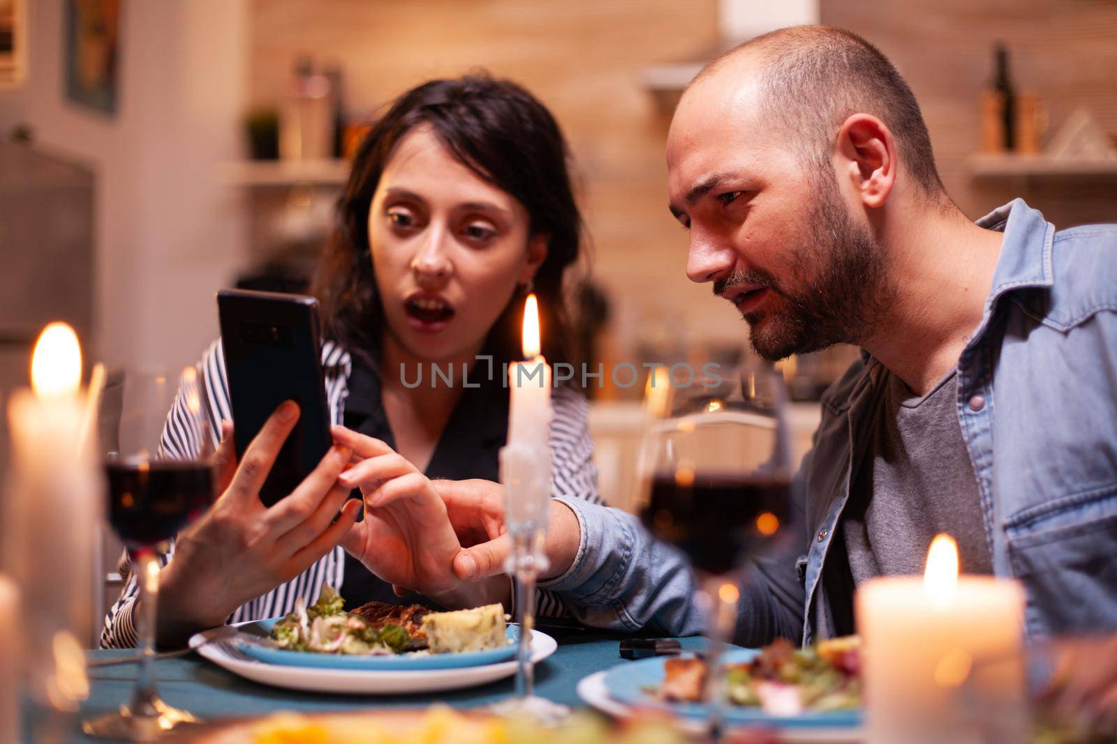 Husband and wife looking shocked at phone while having romantic dinner in kitchen Adults sitting at the table in the kitchen browsing, searching, using smartphones, internet, celebrating anniversary.