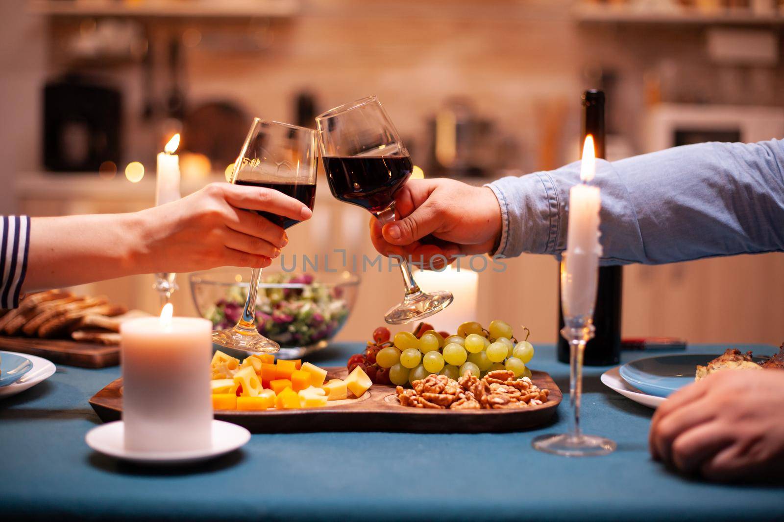 Celebrating anniversary of young couple in kitchen clinking glasses of red wine. Happy cheerful young couple dining together in the cozy kitchen, enjoying the meal.