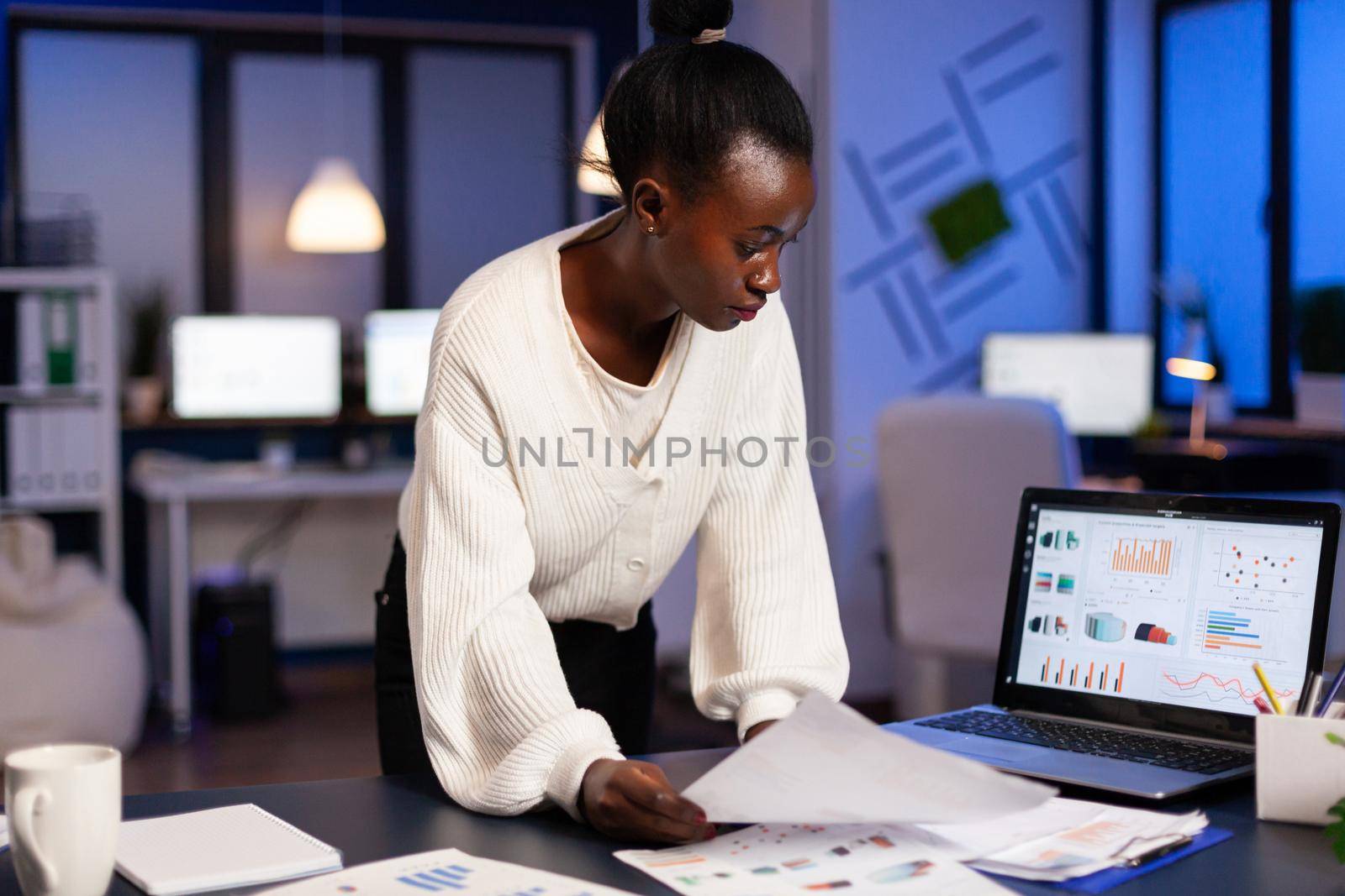 Stressed african manager woman working with financial documents checking graphs, holding papers, reading raports, standing at desk late at night in start-up office doing overtime to respect deadline.