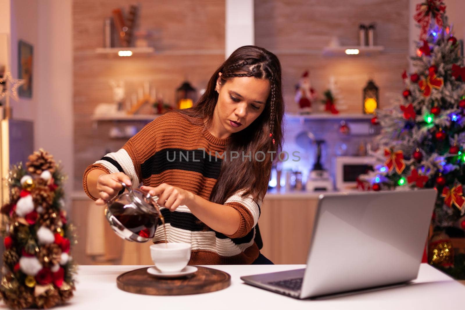 Caucasian woman pouring cup of tea from kettle while talking to friends on video call app. Young adult using laptop for christmas eve in holiday decorated kitchen with ornaments