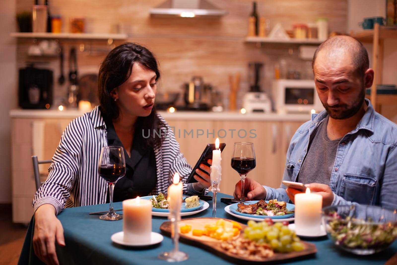 Happy couple watching video on smartphone in kitchen during anniversary. Adults sitting at the table in the kitchen browsing, searching, using smartphones, internet, celebrating anniversary.