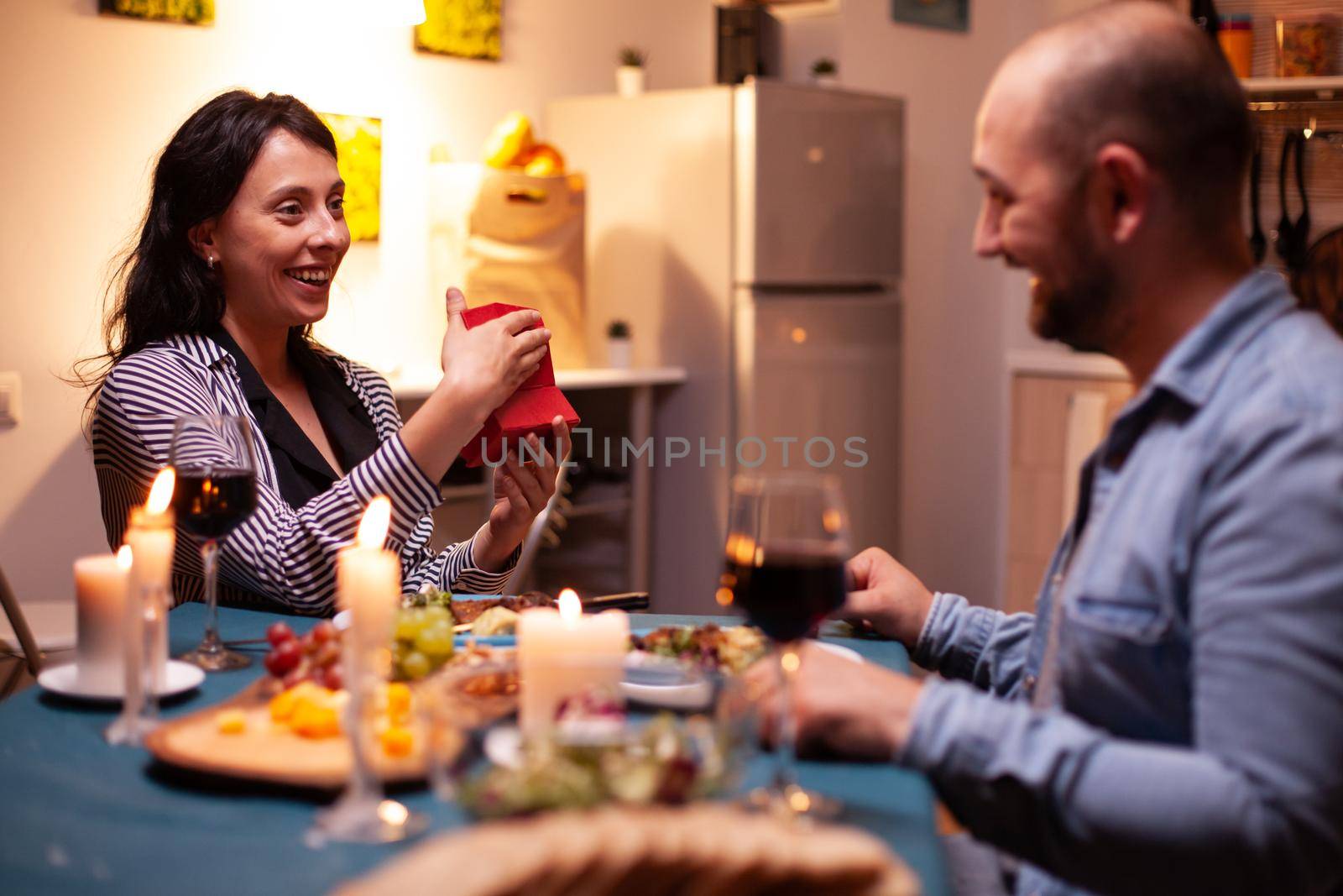 Woman holding anniversary gift from husband during dinner. Happy cheerful couple dining together at home, enjoying the meal celebrating their anniversary.