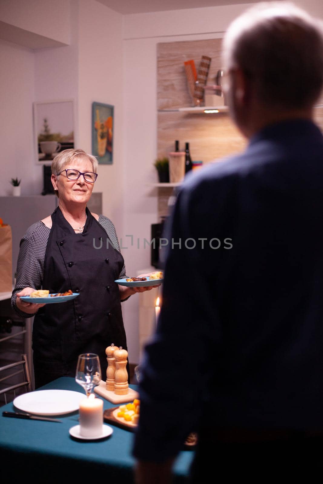 Senior couple having a conversation in kitchen. Elderly aged woman wearing apron. Elderly old couple talking, sitting at the table in kitchen, enjoying the meal, celebrating their anniversary