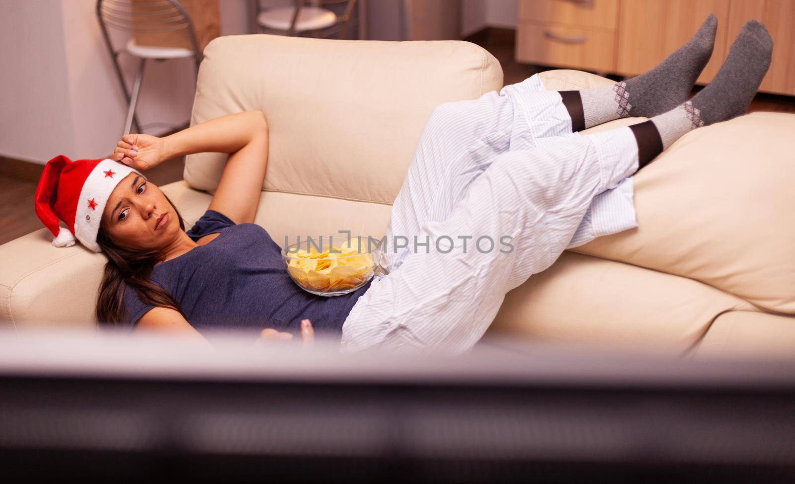 Relaxed girl lying on sofa watching christmas movie series on television celebrating christmastime in x-mas decorated kitchen. Woman wearing santa hat enjoying winter season at home
