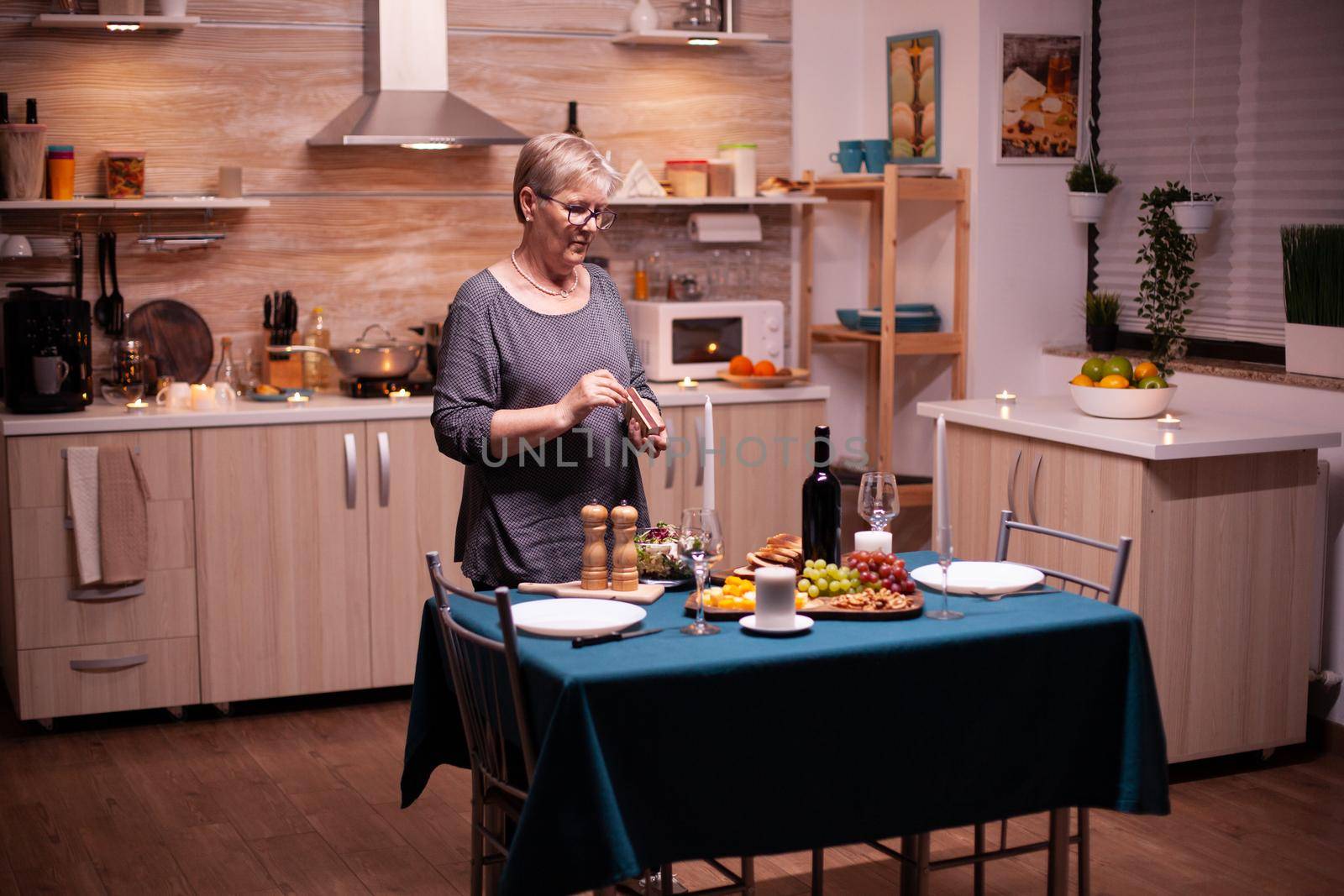 Senior woman lighting candles in kitchen for romantic dinner with husband. Elderly woman waiting her husband for a romantic dinner. Mature wife preparing festive meal for anniversary celebration.