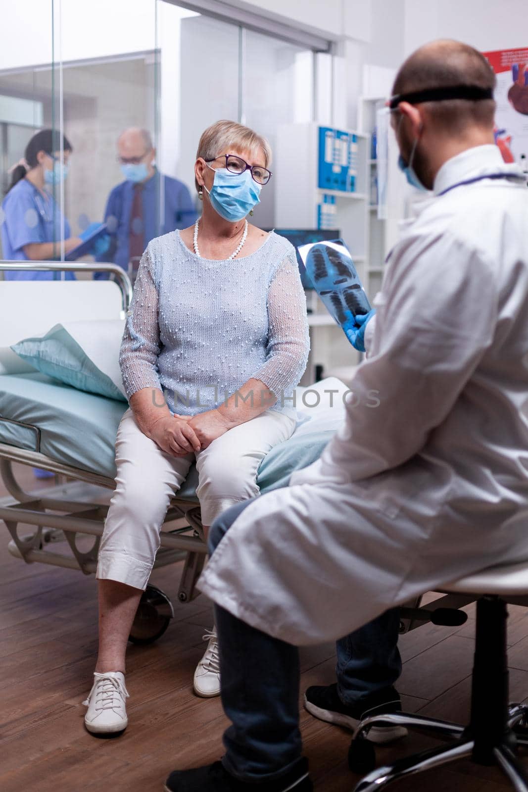 Medical specialist in hospital room examining x-ray of old woman and giving a diagnosis while wearing protection mask against coronavirus as safety precaution. Check up for infections, disease .