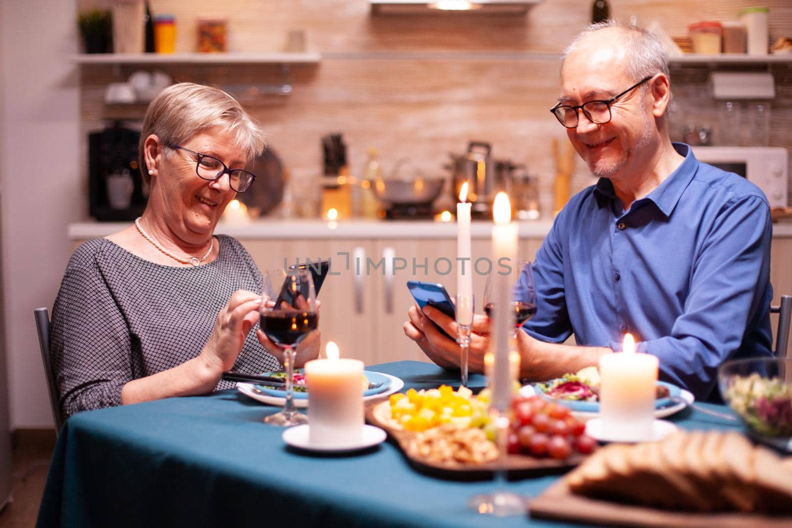 Retired man browsing on smartphone in kitchen and having dinner with wife. Sitting at the table in the kitchen, browsing, searching, using phone, internet, celebrating their anniversary in the dining room.