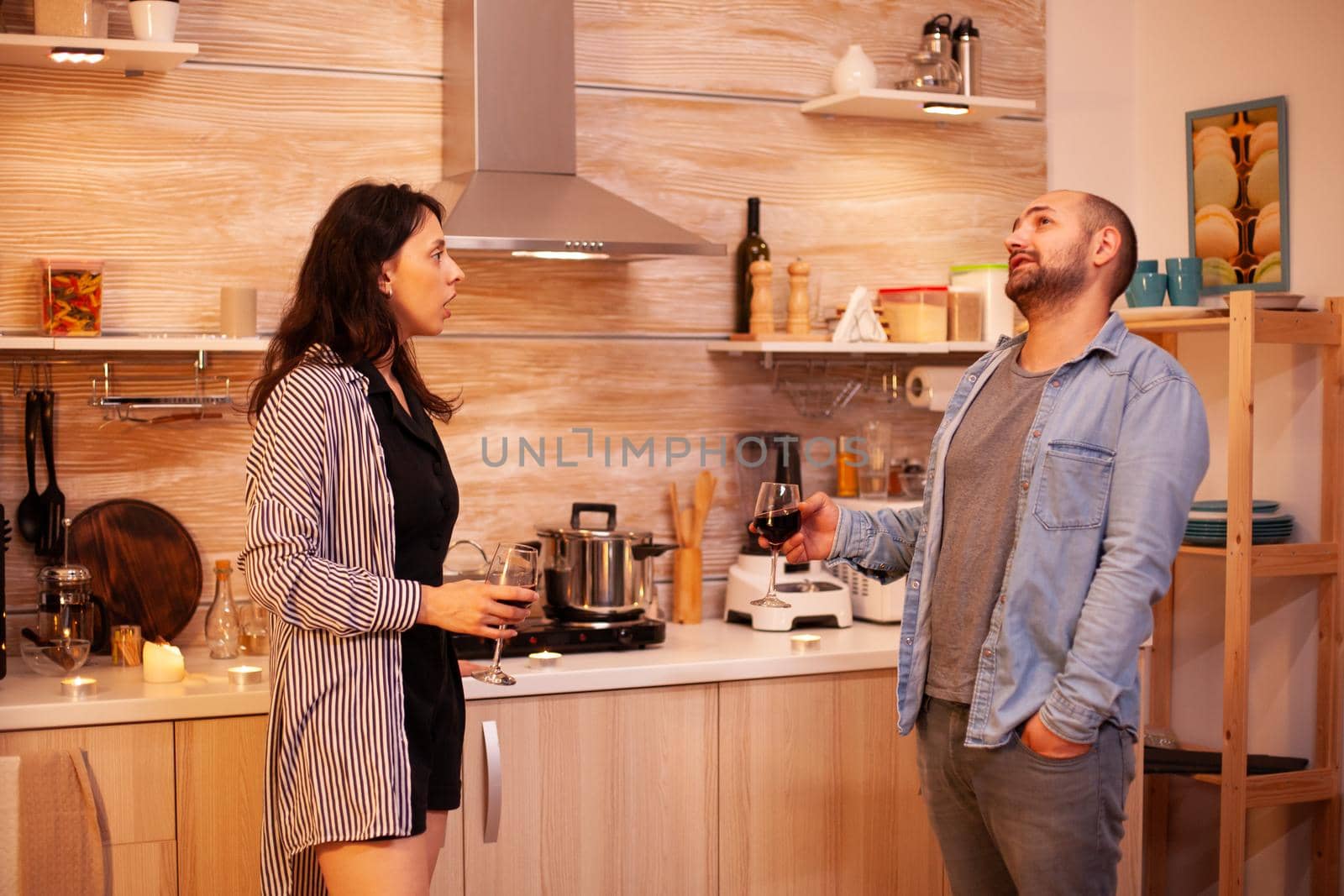 Wife telling a story to her husband while holding a glass of wine in kitchen. Adult couple at home, drinking red wine, talking, smiling, enjoying the meal in dining room.
