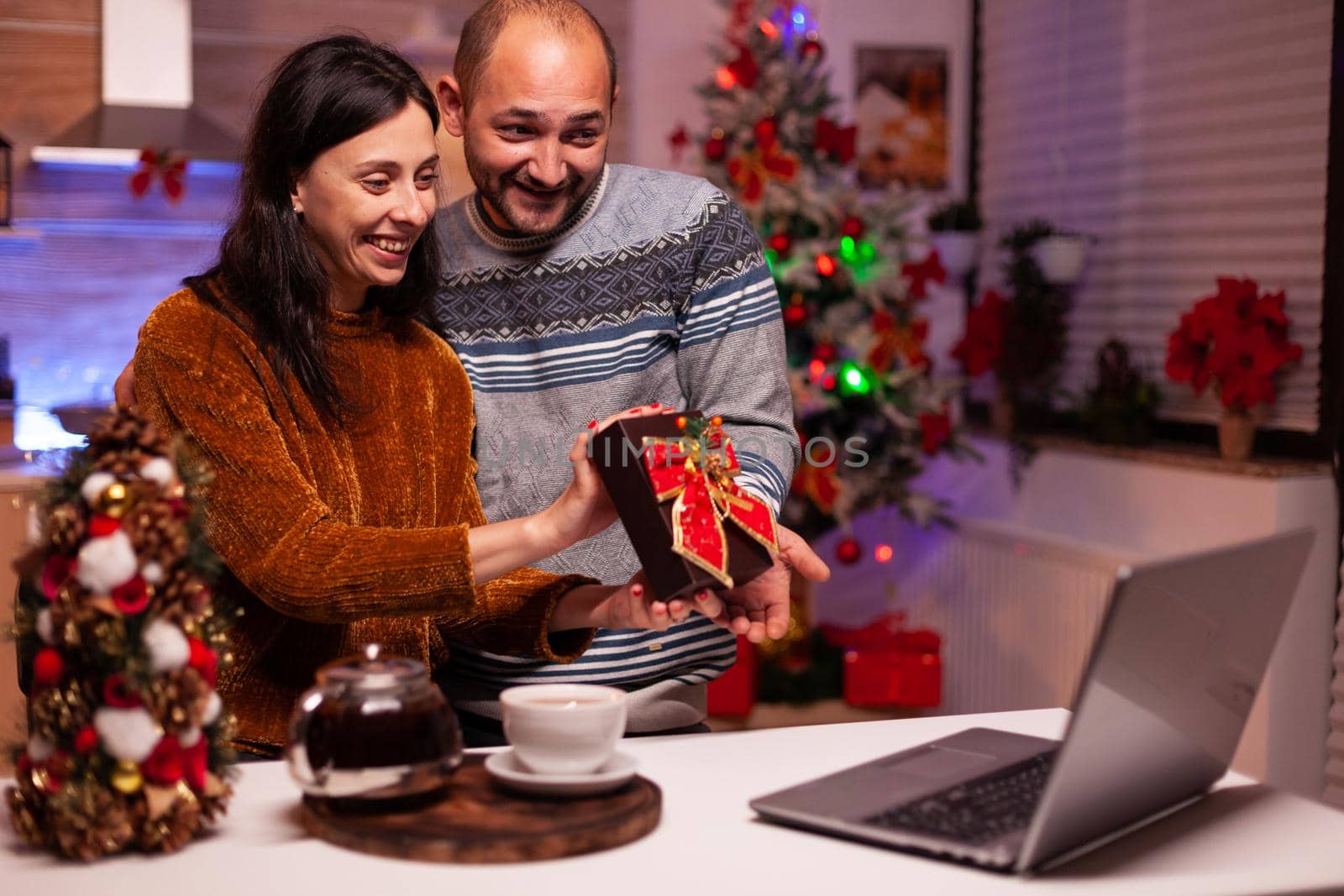 Happy family showing xmas present surprise to remote friends during online videocall meeting on laptop computer standing in festive decorated kitchen. Cheerful couple enjoying christmas holiday