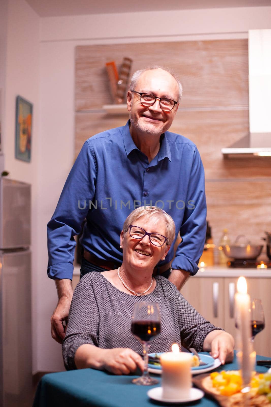 Excited disabled woman in wheelchair and her husband behind her looking at camera. Happy cheerful senior elderly couple dining together in the cozy kitchen, enjoying the meal, celebrating their anniversary.