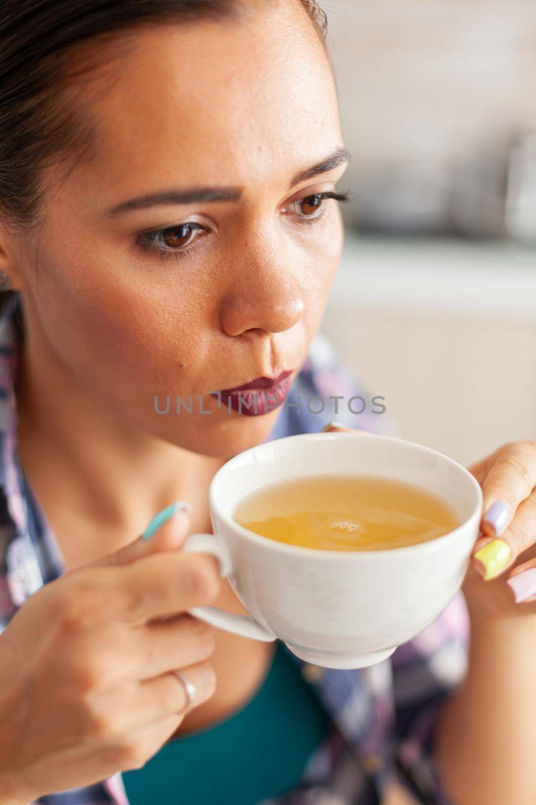 Woman trying to drink hot green tea with aromatic herbs in kitchen in the morning. Pretty lady sitting in the kitchen in the morning during breakfast time relaxing with tasty natural herbal tea.