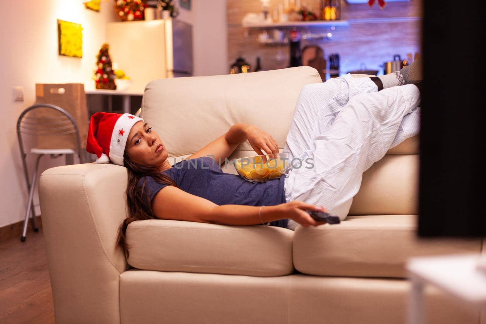 Woman resting on couch eating snacks while searching winter movie on television using remote control enjoying december season. Caucasian female celebrating christmas holiday in xmas decorated kitchen