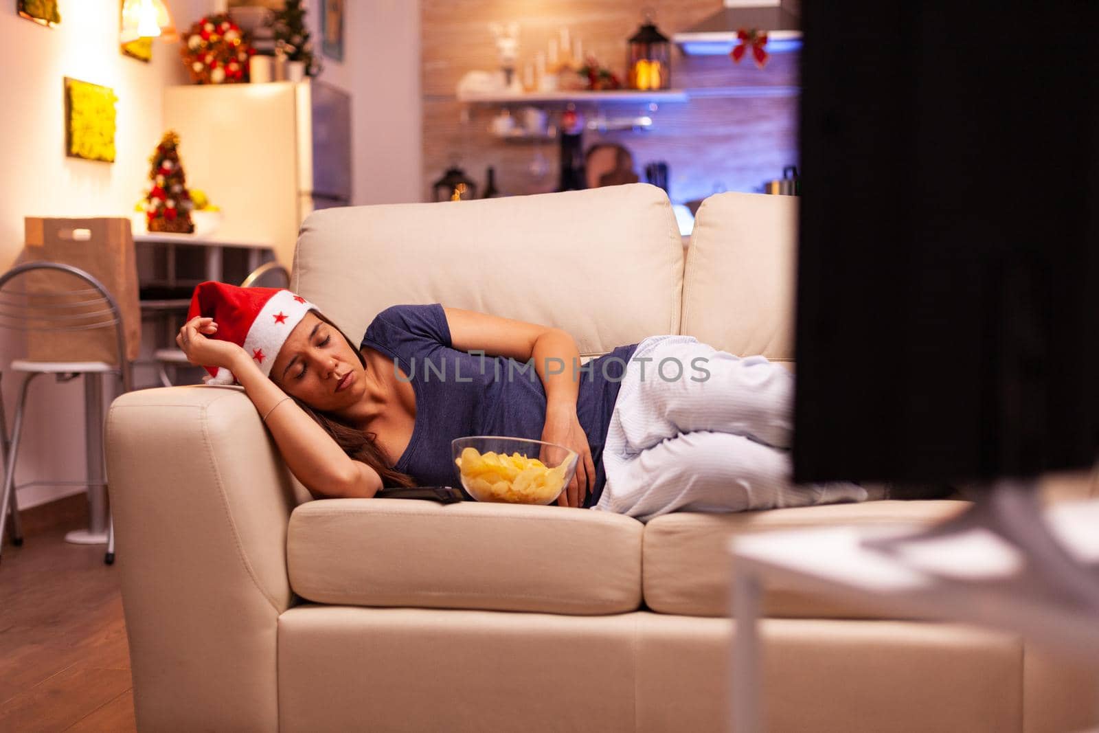 Girl falling asleep on sofa in xmas decorated kitchen after watching winter movie on television. Caucasian female with red santa hat enjoying christmas holiday. X-mas season