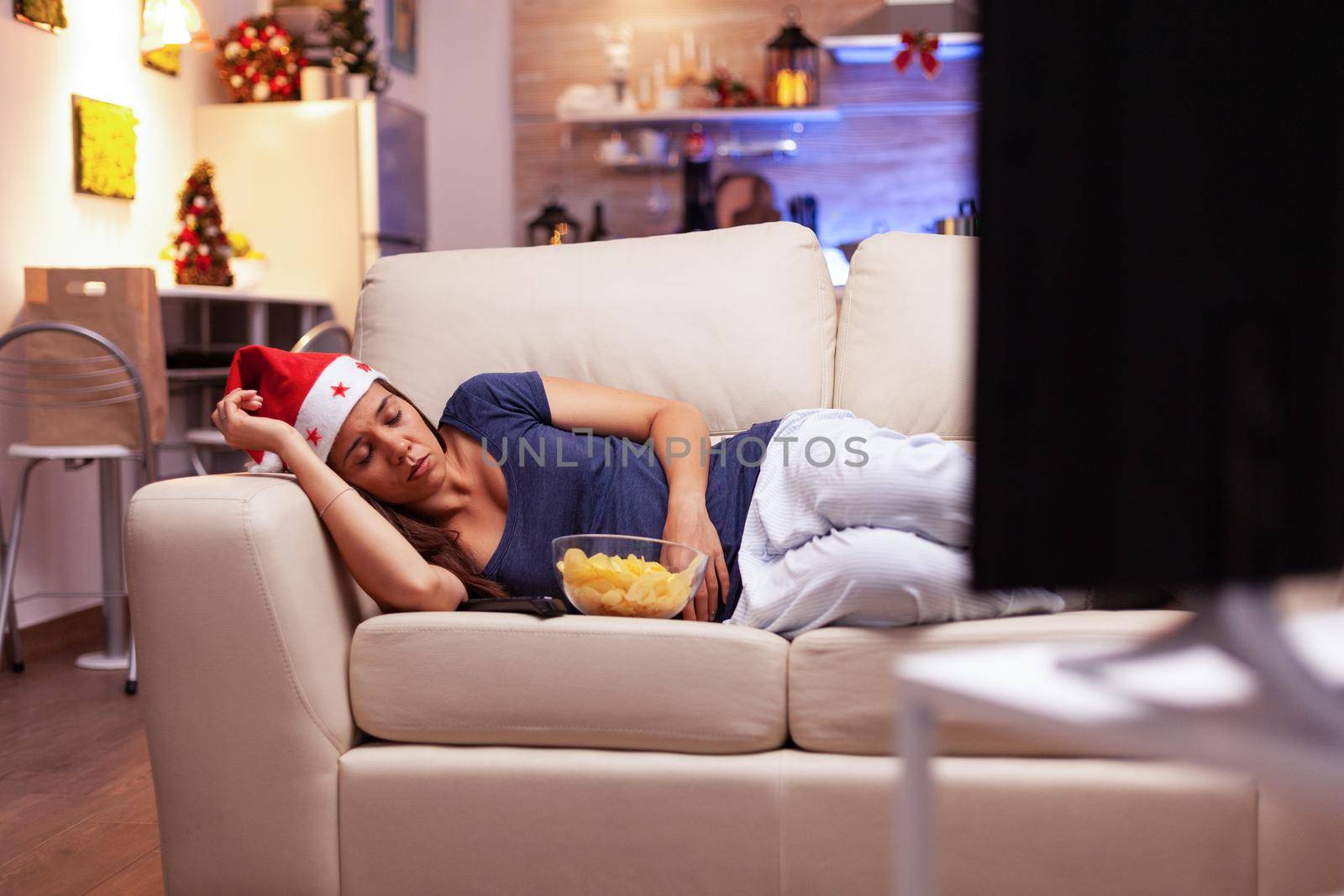 Tired woman falling asleep on sofa while watching xmas movie on television during christmastime enjoying winter season in x-mas decorated kitchen. Adult person celebrating christmas holiday