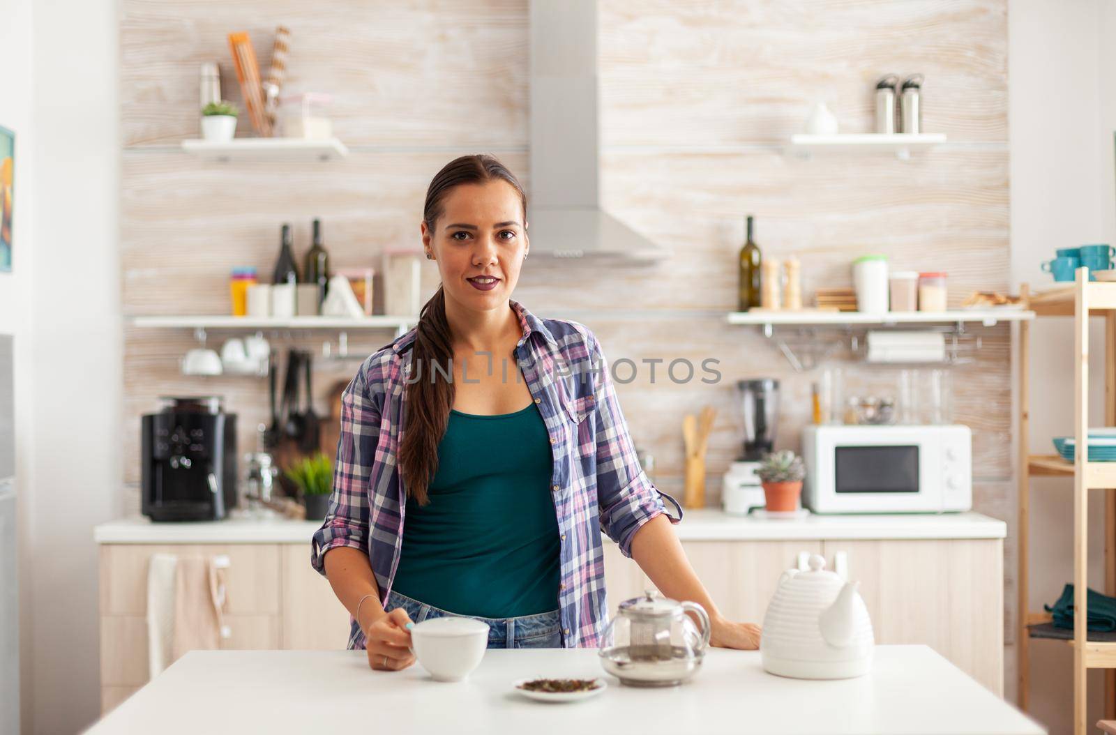 Woman holding cup of tea during morning in kitchen while enjoying breakfast. Preparing tea in the morning, in a modern kitchen sitting near the table. Putting with hands, healthy herbal in pot.