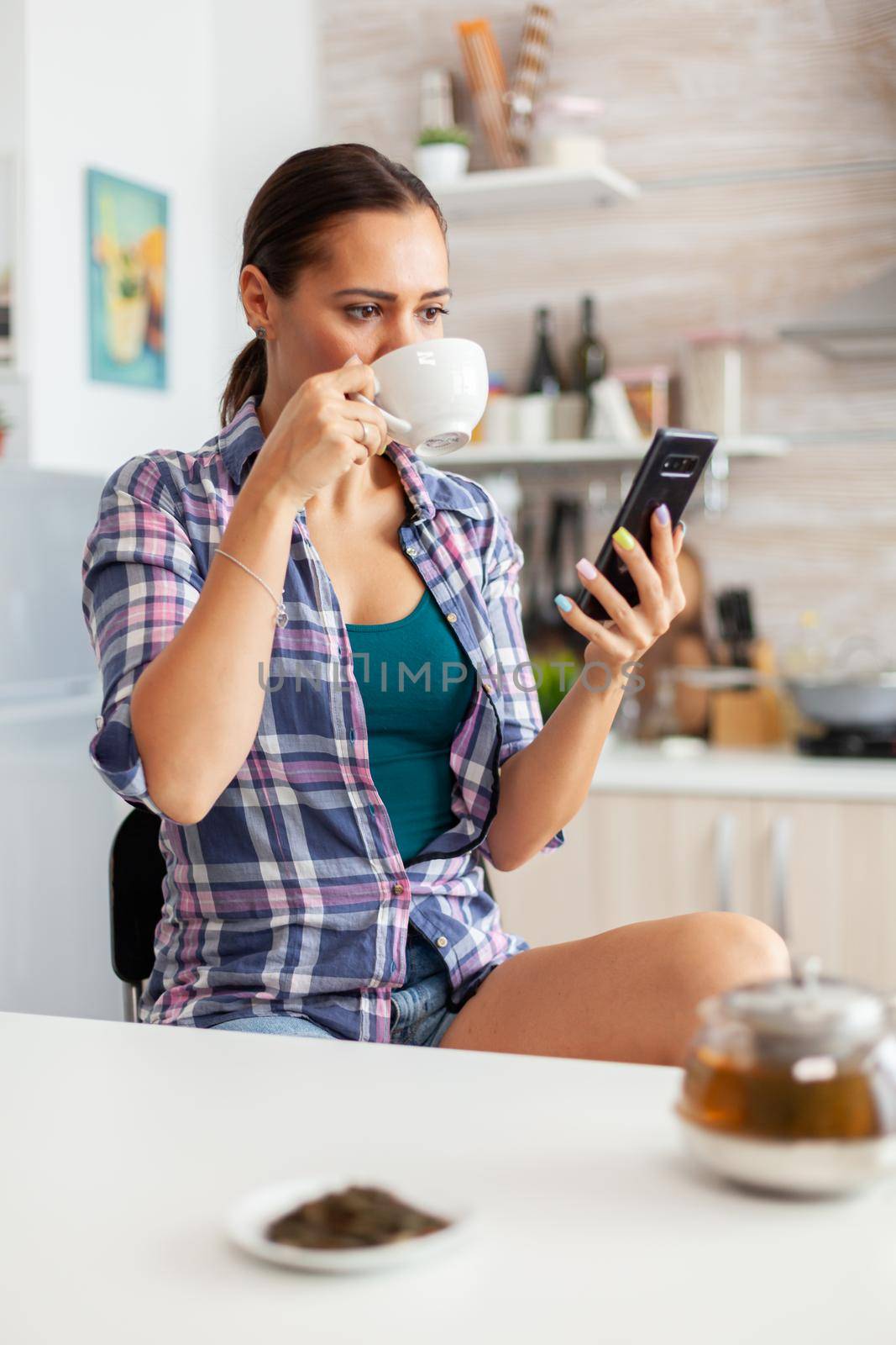 Housewife drinking hot green tea using smartphone in the morning during breakfast. Holding phone device with touchscreen using internet technology scrolling, searching on intelligent gadget.