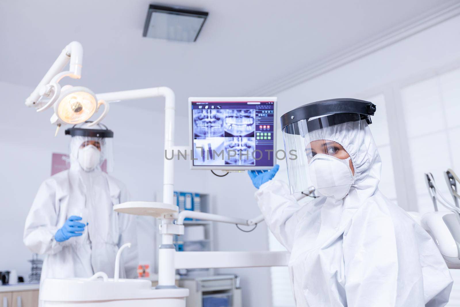 Patient pov listening explication for teeth treatment showing x-ray on monitor. Stomatology specialist wearing protective suit against infection with coronavirus pointing at radiography.