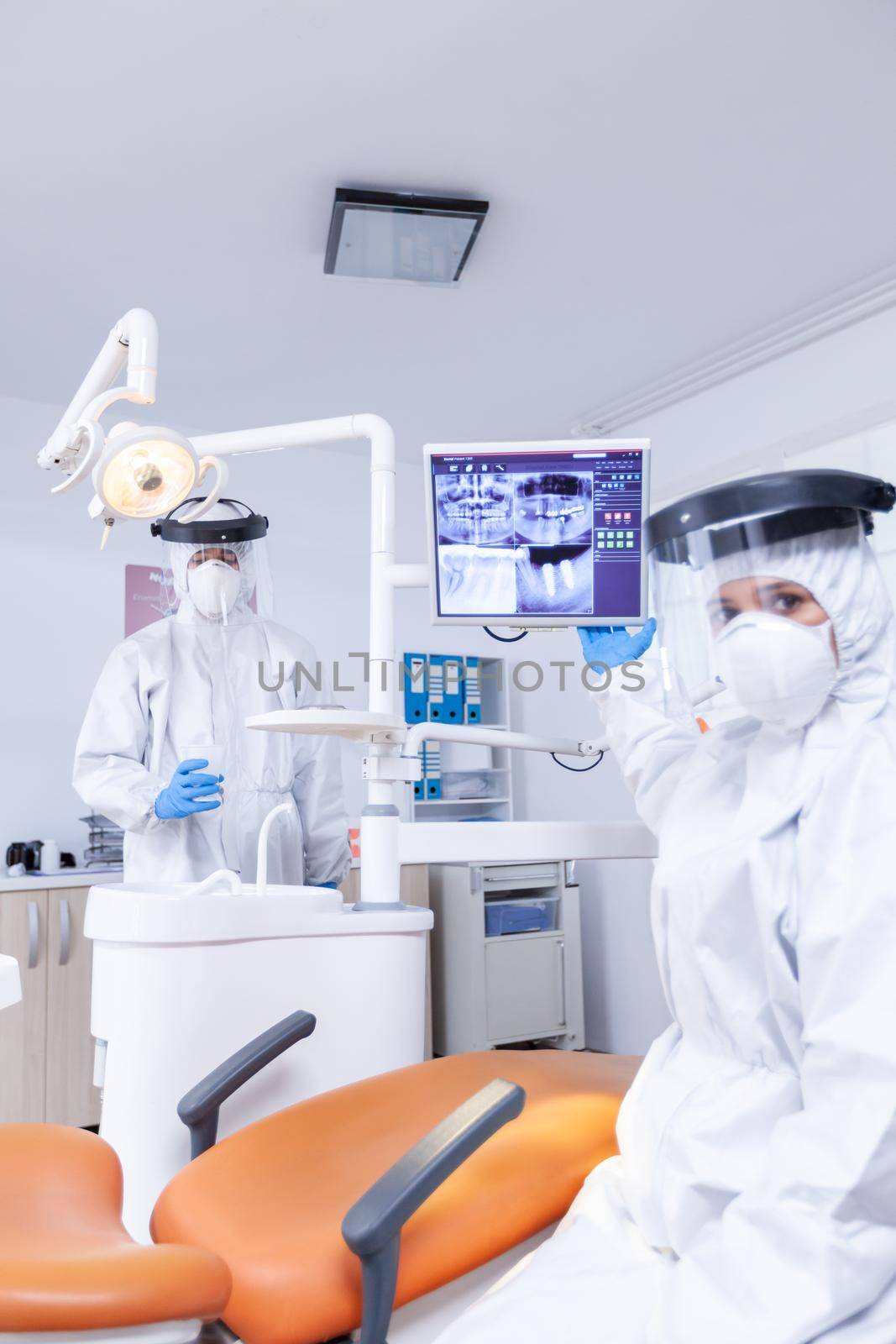 Patient pov of dentist explaining radiography of teeth from digital monitor. Stomatology specialist wearing protective suit against infection with coronavirus pointing at radiography.