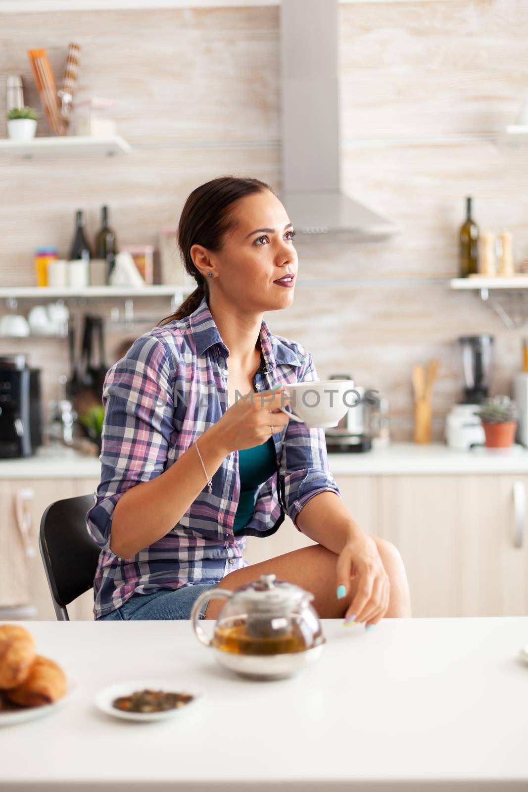 Dreamy girl enjoying hot green tea during breakfast in kitchen. Young lady relaxing with tea in the morning sitting in home enjoying pleasant memories.