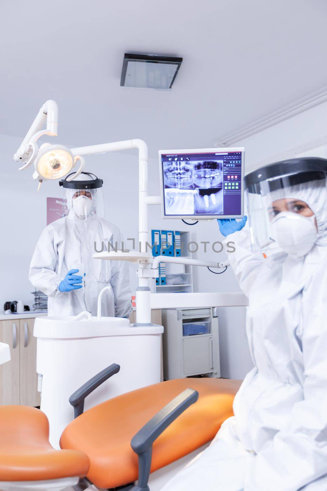 Stomatologist telling the treatment to patient showing digital teeth x-ray. Stomatology specialist wearing protective suit against infection with coronavirus pointing at radiography.