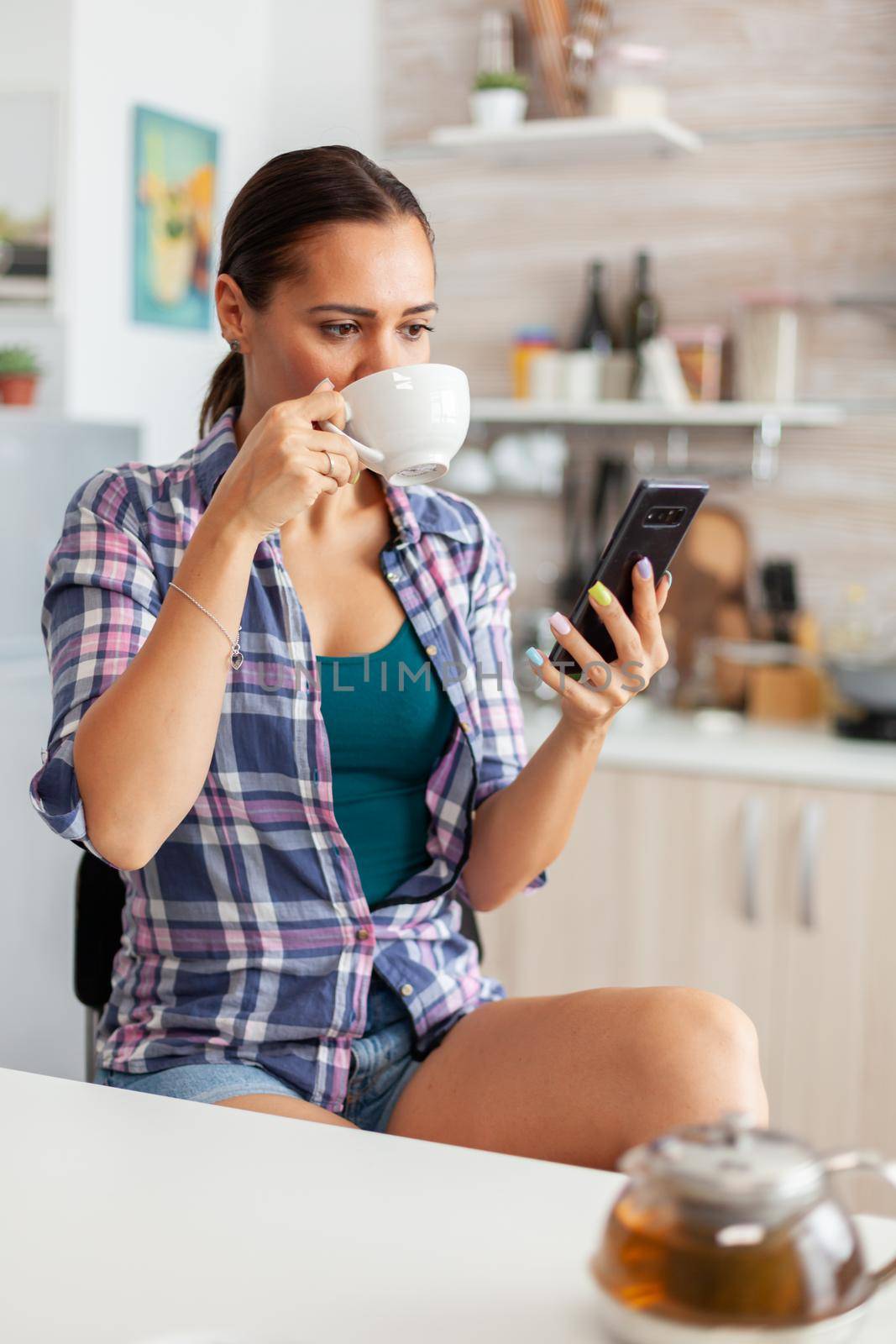 Woman drinking aromatic tea in the morning browsing on smartphone. Holding phone device with touchscreen using internet technology scrolling, searching on intelligent gadget.
