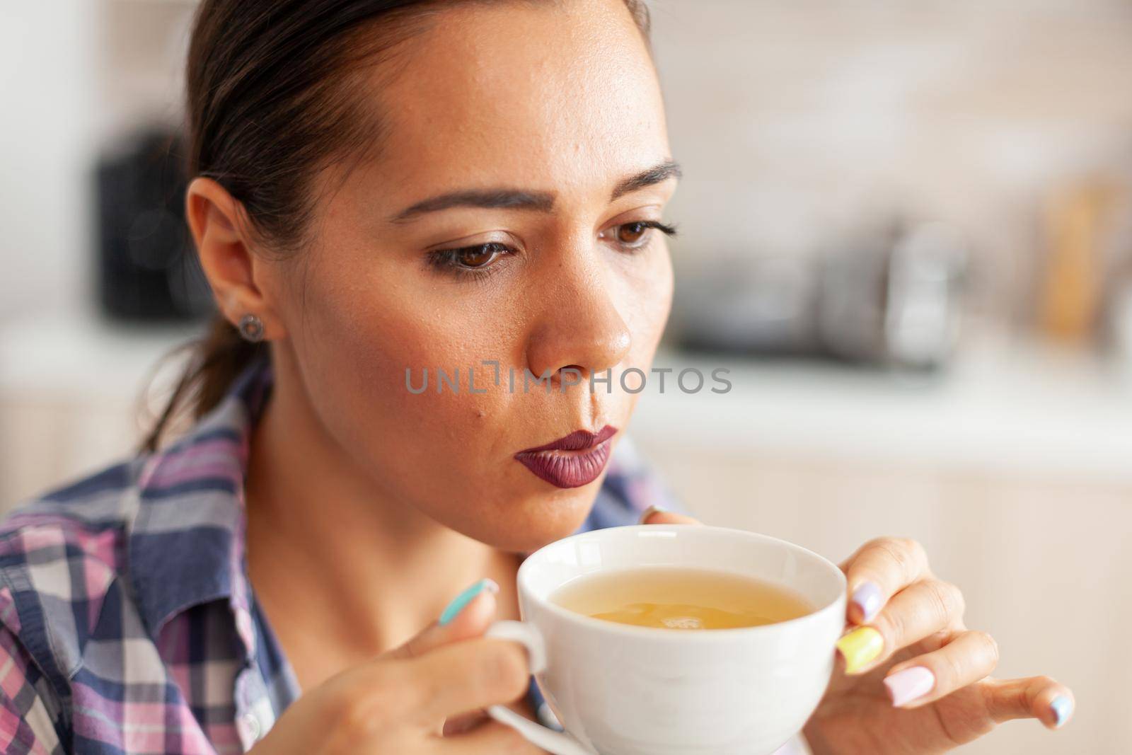 Close up of woman in kitchen trying to drink hot green tea with aromatic herbs. Pretty lady sitting in the kitchen in the morning during breakfast time relaxing with tasty natural herbal tea.