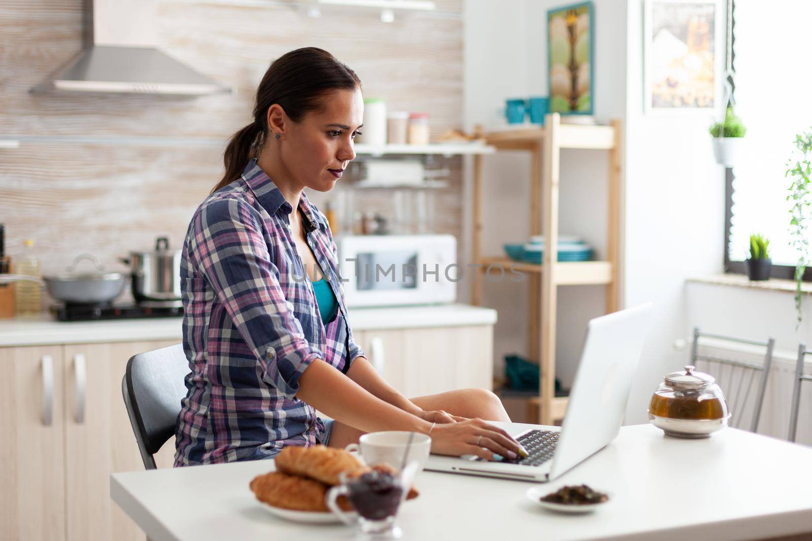 Woman working from home using laptop in kitchen during breakfast. Working from home using device with internet technology, browsing, searching on gadget in the morning.