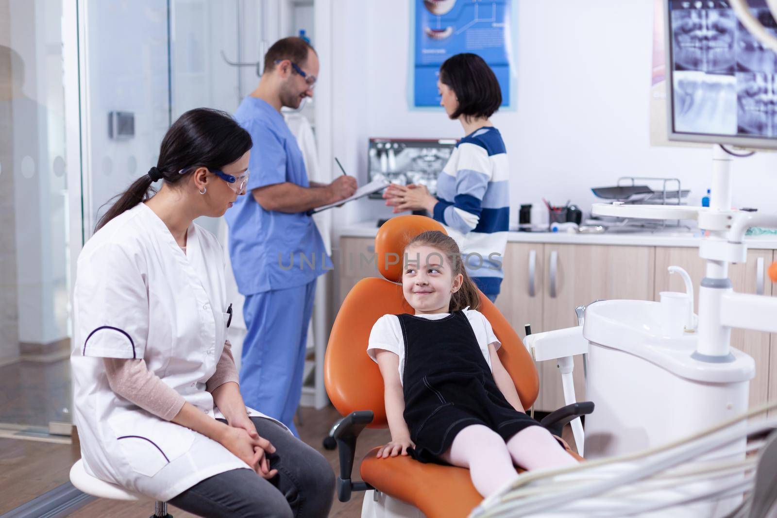 Shy little girl looking up at dentist doctor waiting teeth cavity treatment. Child with her mother during teeth check up with stomatolog sitting on chair.
