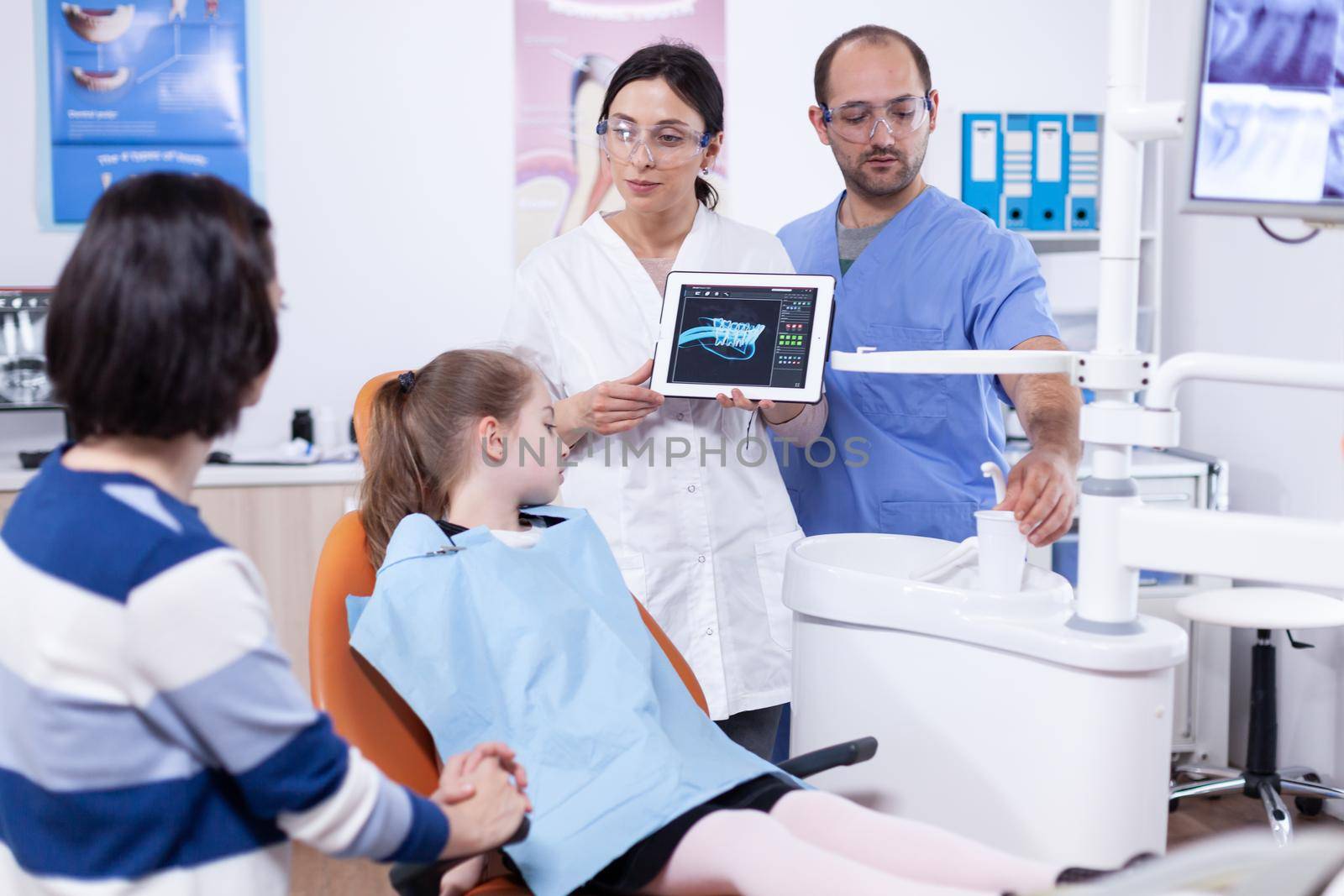 Dentist in dentistiry office showing tablet pc with jaw radiography to parent and little girl explaining treatment. Stomatologist explaining teeth diagnosis to mother of child in health clinic holding x-ray.