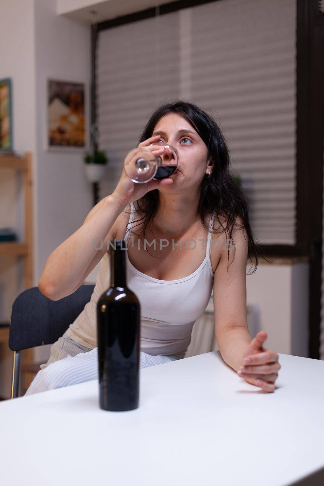 Alcoholic woman having bottle of wine and glass feeling sad at home. Lonely person drinking beverage with alcohol being depressive. Adult with addiction feeling emotional and upset