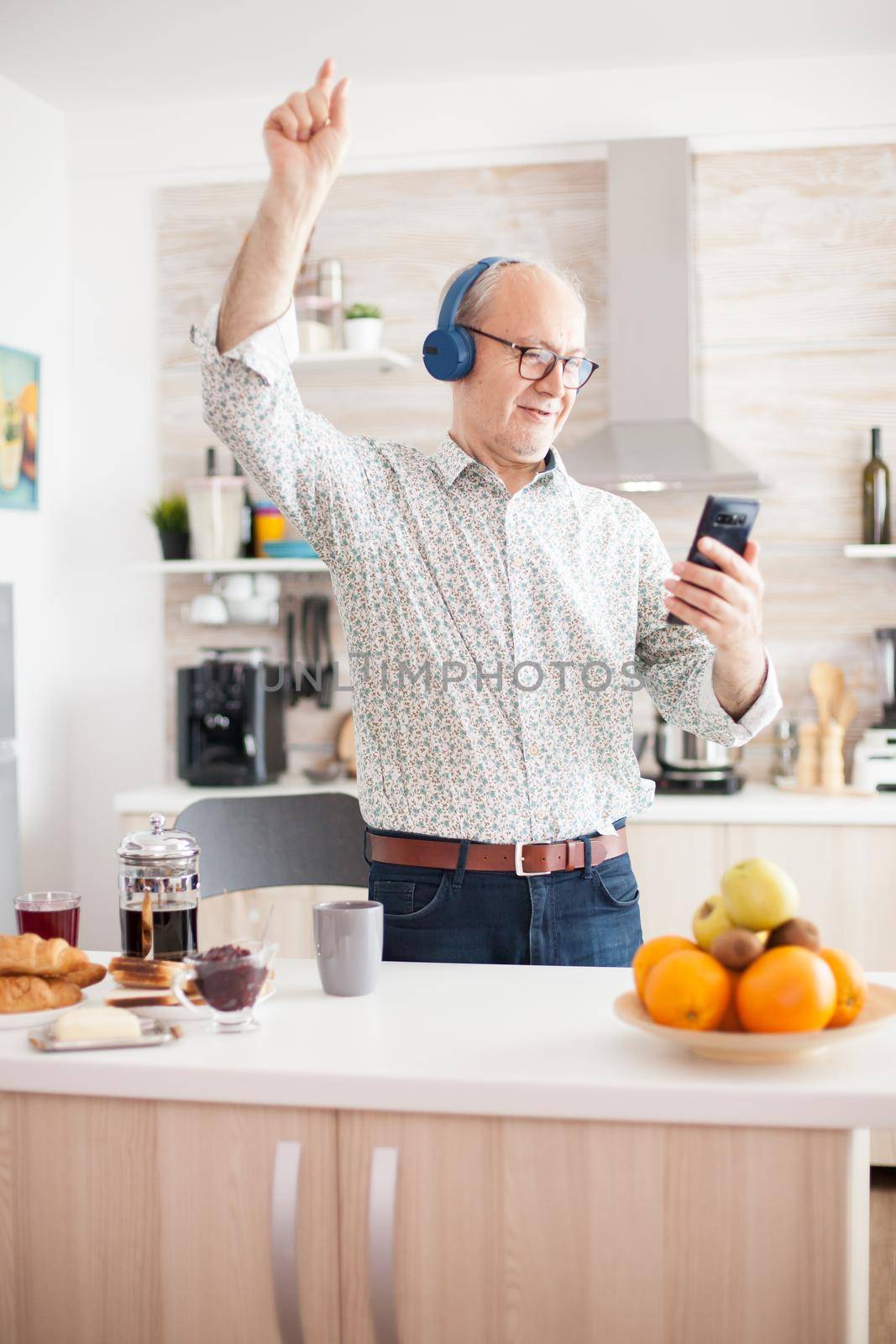 Happy senior man listening music wearing headphones in kitchen during breakfast. Elderly retired person enjoying modern fun happy lifestyle, dancing relaxed, smiling and using technology