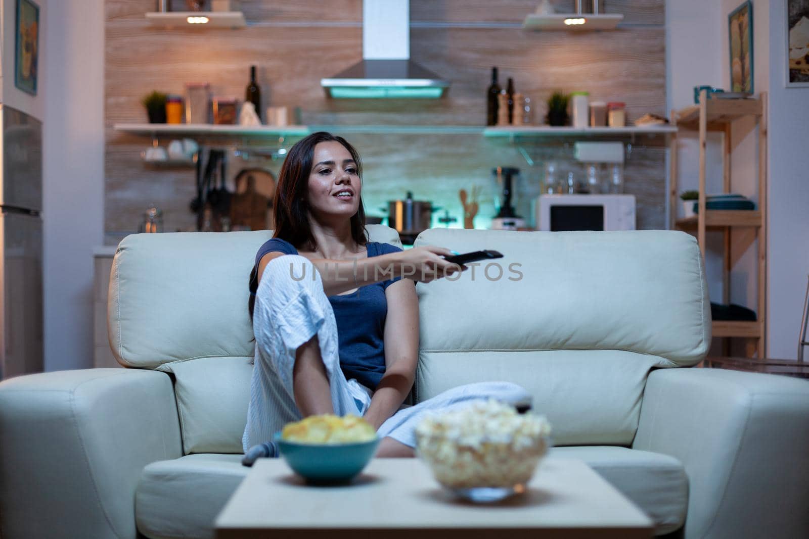 Caucasian woman sitting on couch and watching tv relaxing after work. Excited amused home alone lady in pijamas resting with snacks and juice sitting on comfortable sofa in open space living room.