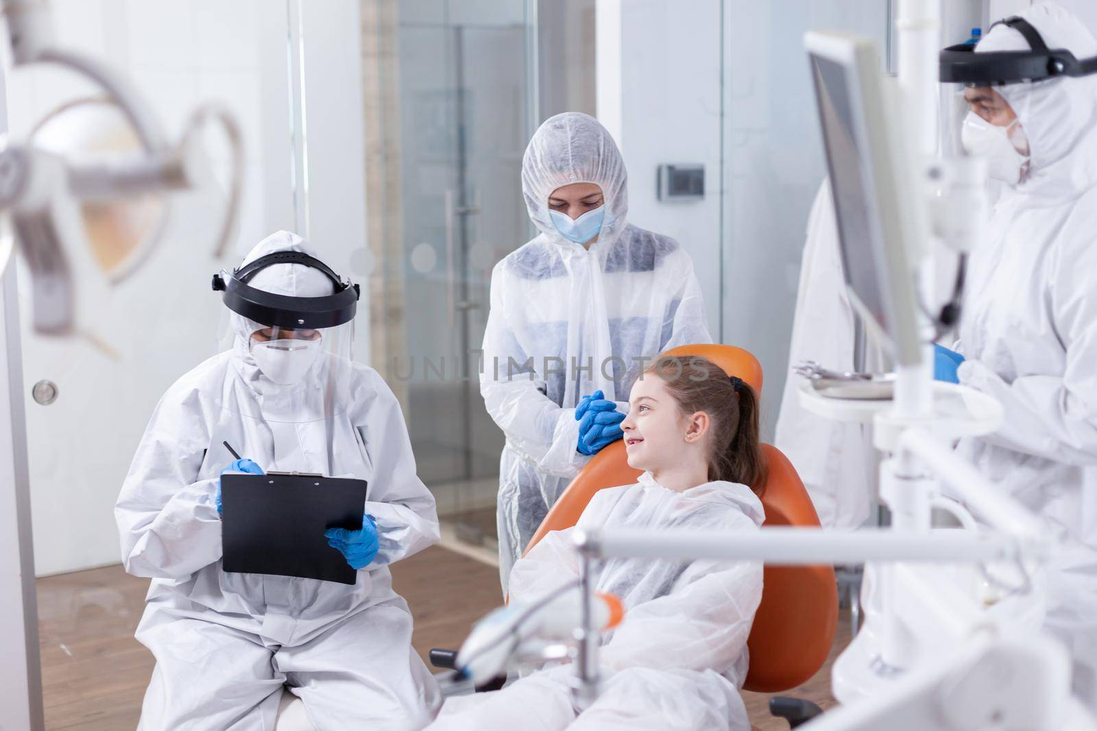 Little girl smiling at pediatric dentist sitting on dental chair in the course of dentistiry check up. Stomatologist during covid19 wearing ppe suit doing teeth procedure of child sitting on chair.