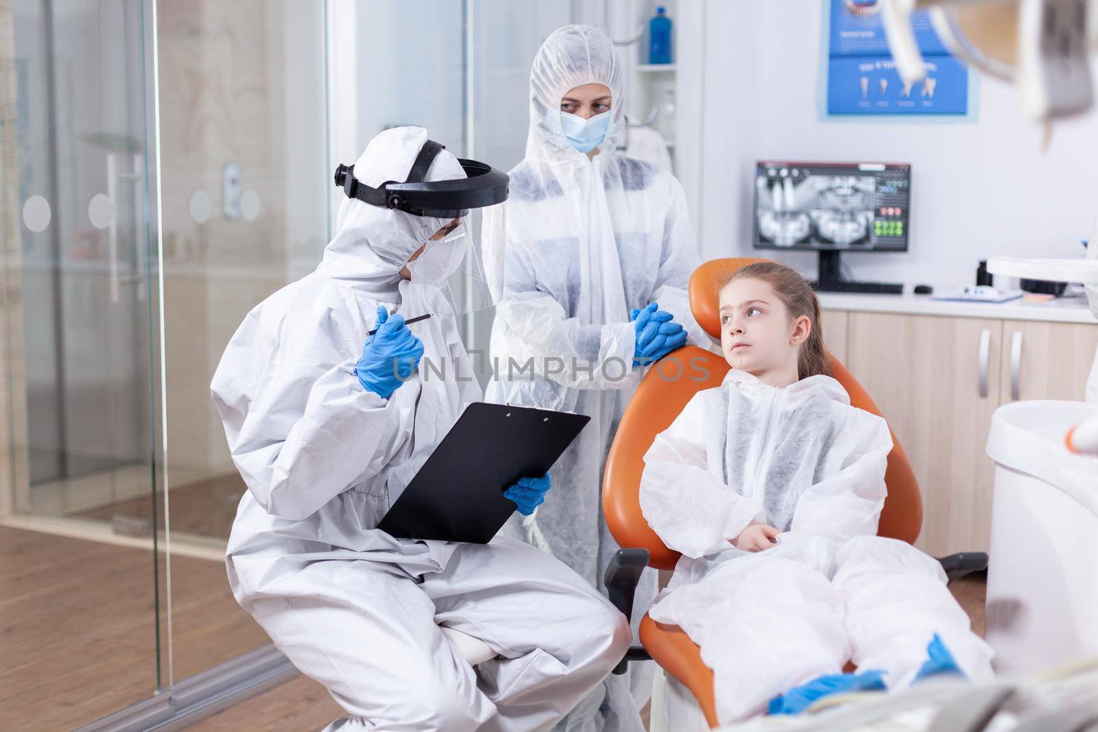 Orthodontist explaining cavity treatment to parent and little girl dressed in protection suit against coronavirus. Stomatologist during covid19 wearing ppe suit doing teeth procedure of child sitting on chair.