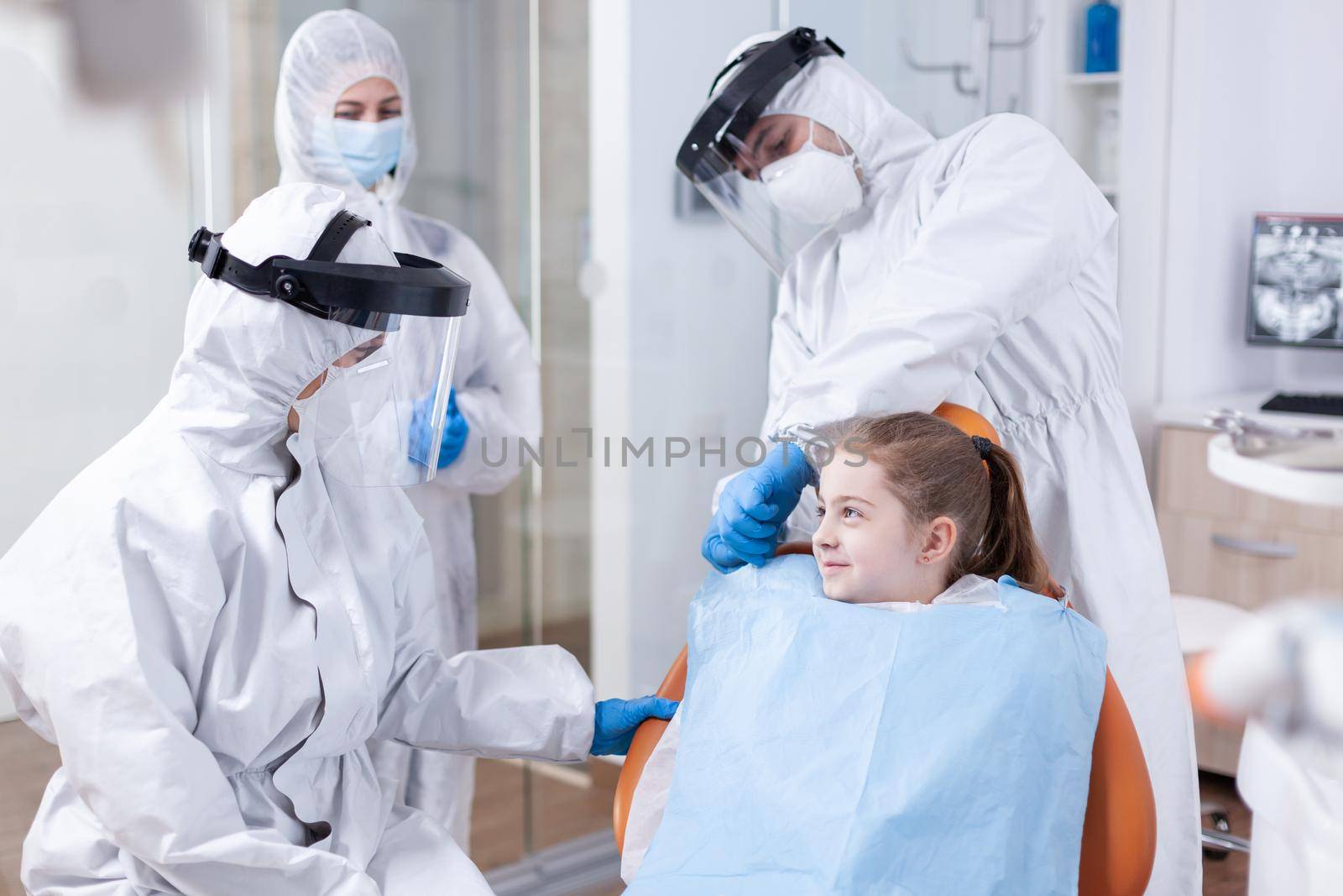 Smilling little girl in the course of dental examination dressed in coverall because of coronavirus pandemic. Stomatologist during covid19 wearing ppe suit doing teeth procedure of child sitting on chair.