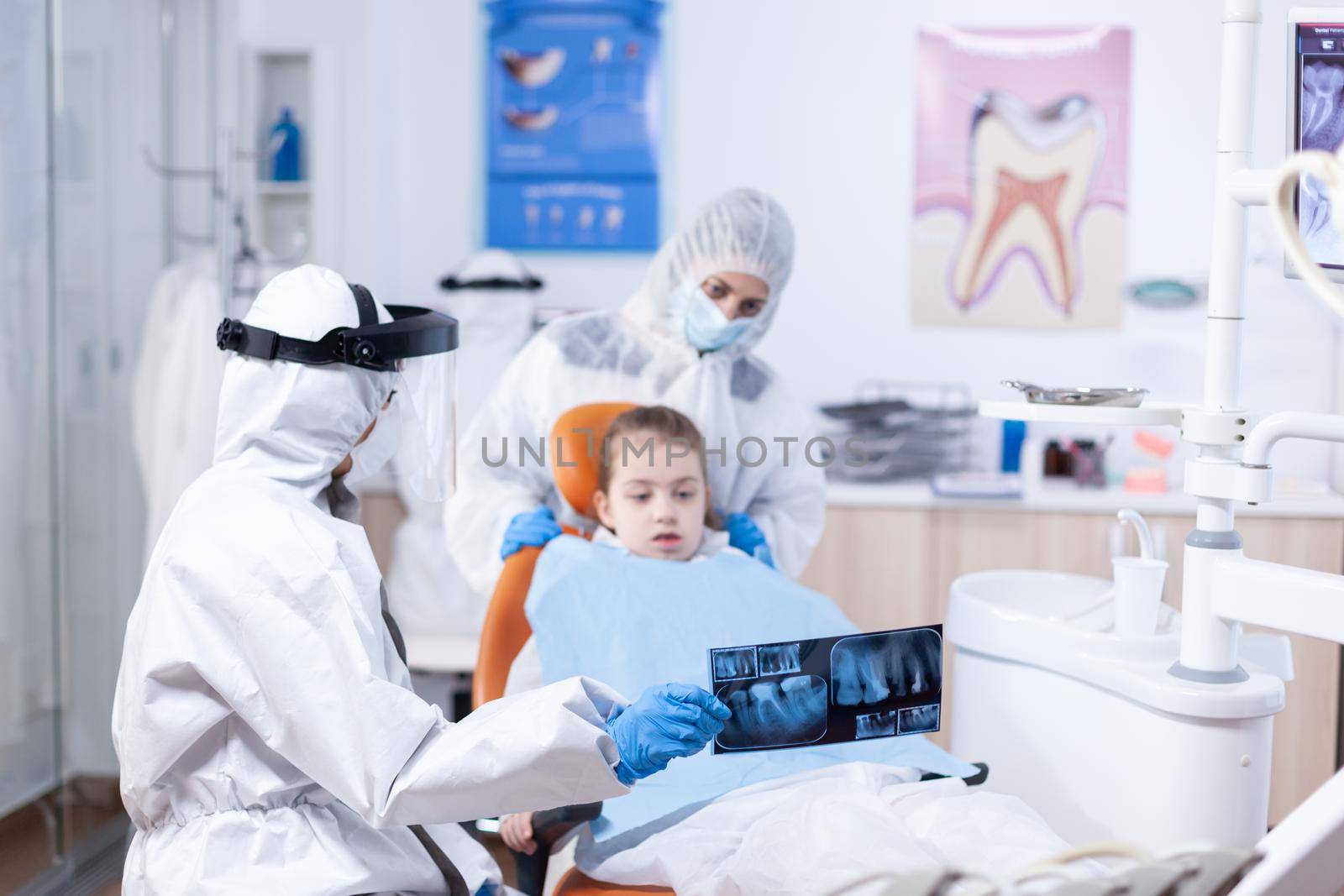 Dentist dressed in ppe suit reviewing panoramic mouth radiography of little girl patient. Stomatolog in protectie suit for coroanvirus as safety precaution holding child teeth x-ray during consultation.