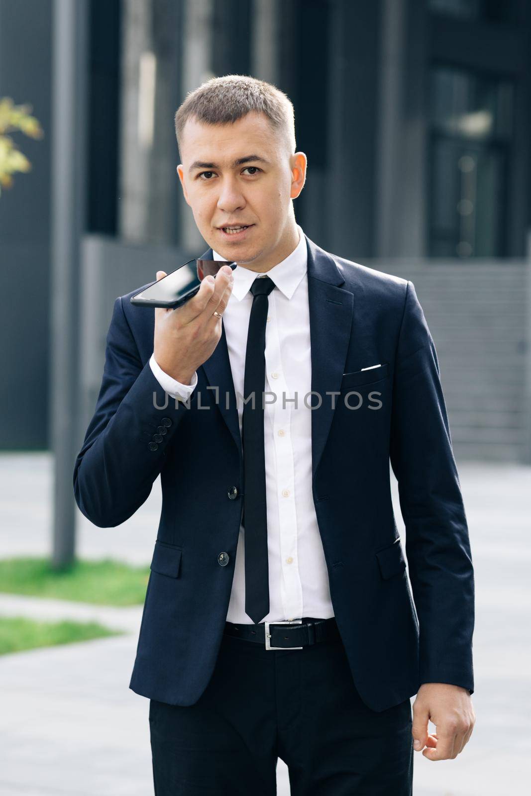 Businessman use smartphone to send voice messages outdoors at downtown. Young business man talking on phone near modern office building.