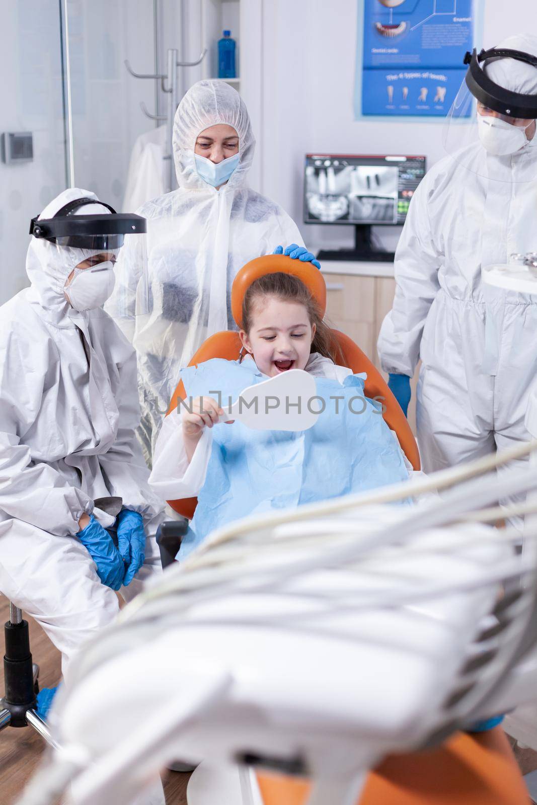 Cheerful little girl looks in mirror after dentral treatment dressed in ppe suit. Child wearing ppe suit during teeth intervention at dental hospital.