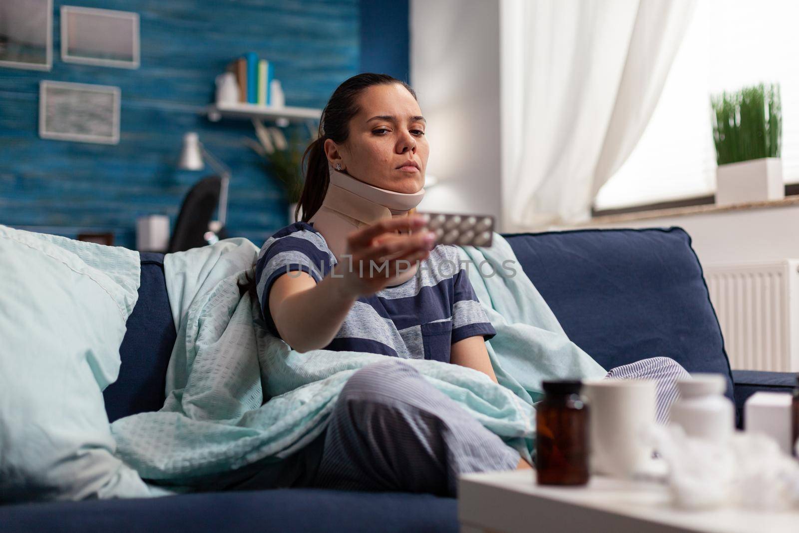 Woman with neck collar suffering from cervical pain sitting on couch taking medical treatment for muscle injury. Young adult with temporary physical joint discomfort on sofa