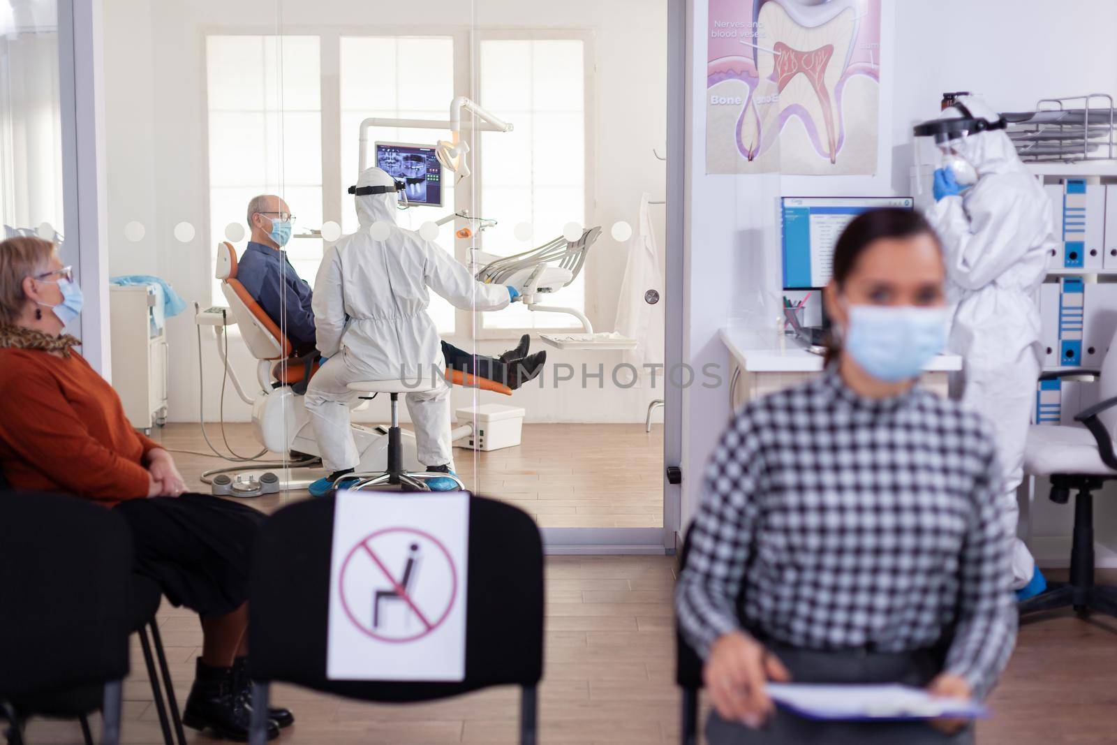 Doctor in pee suit consulting senior patient in stomatology office keeping visor, during global pandemic with coronavirus, new normal. Receptionist with face mask in dentistiry waiting area.